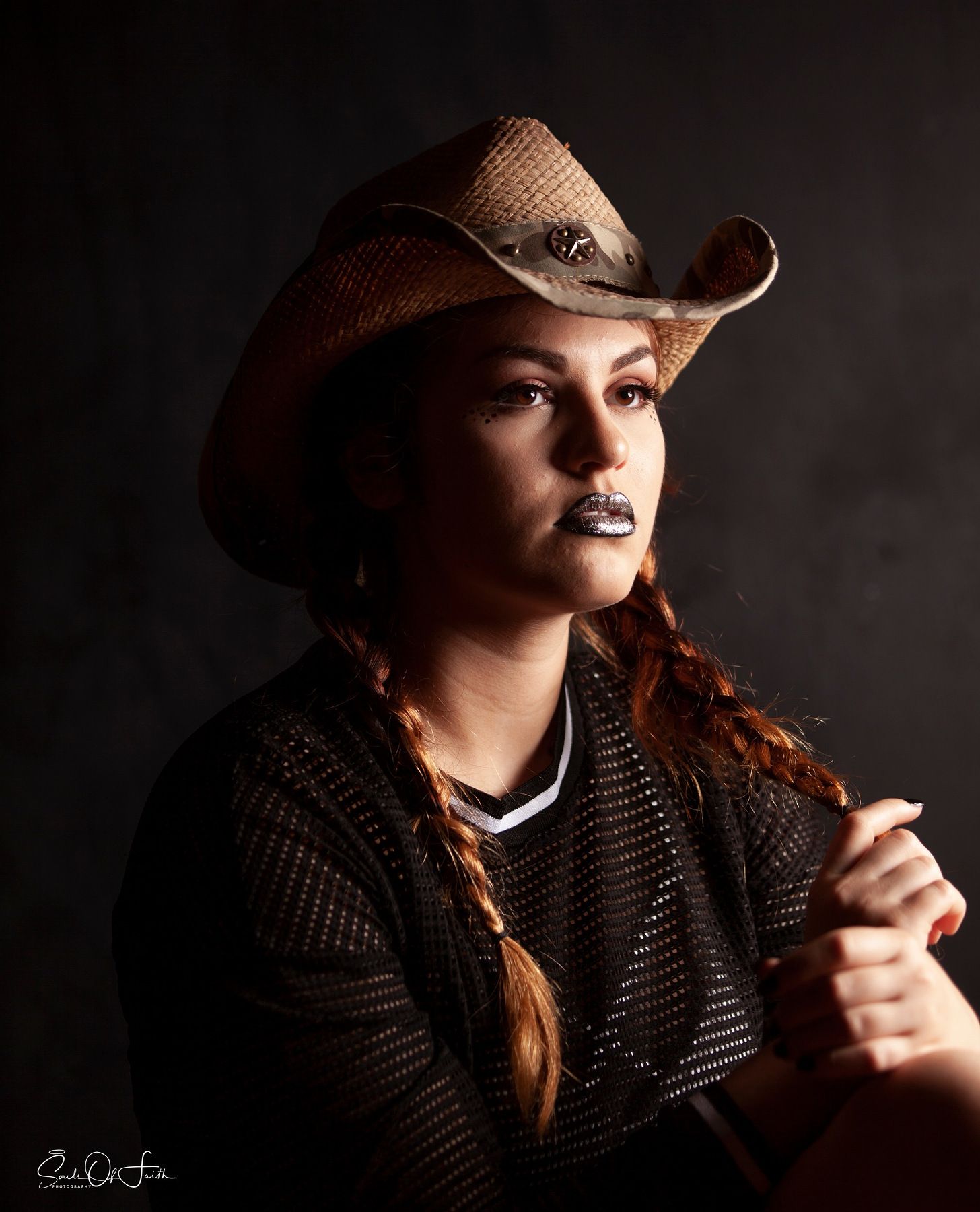 Young woman in straw cowboy hat with braids and dark lipstick poses in dramatic studio lighting.