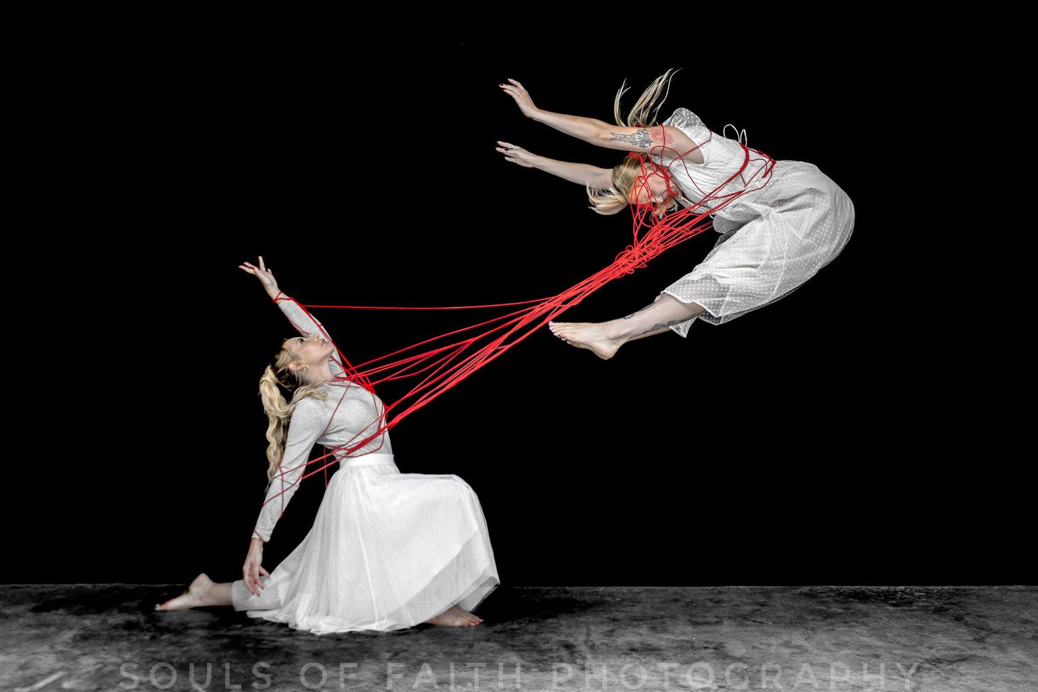 Two dancers in white connected by red ribbons, one seated and one leaping mid-air against a dark background.