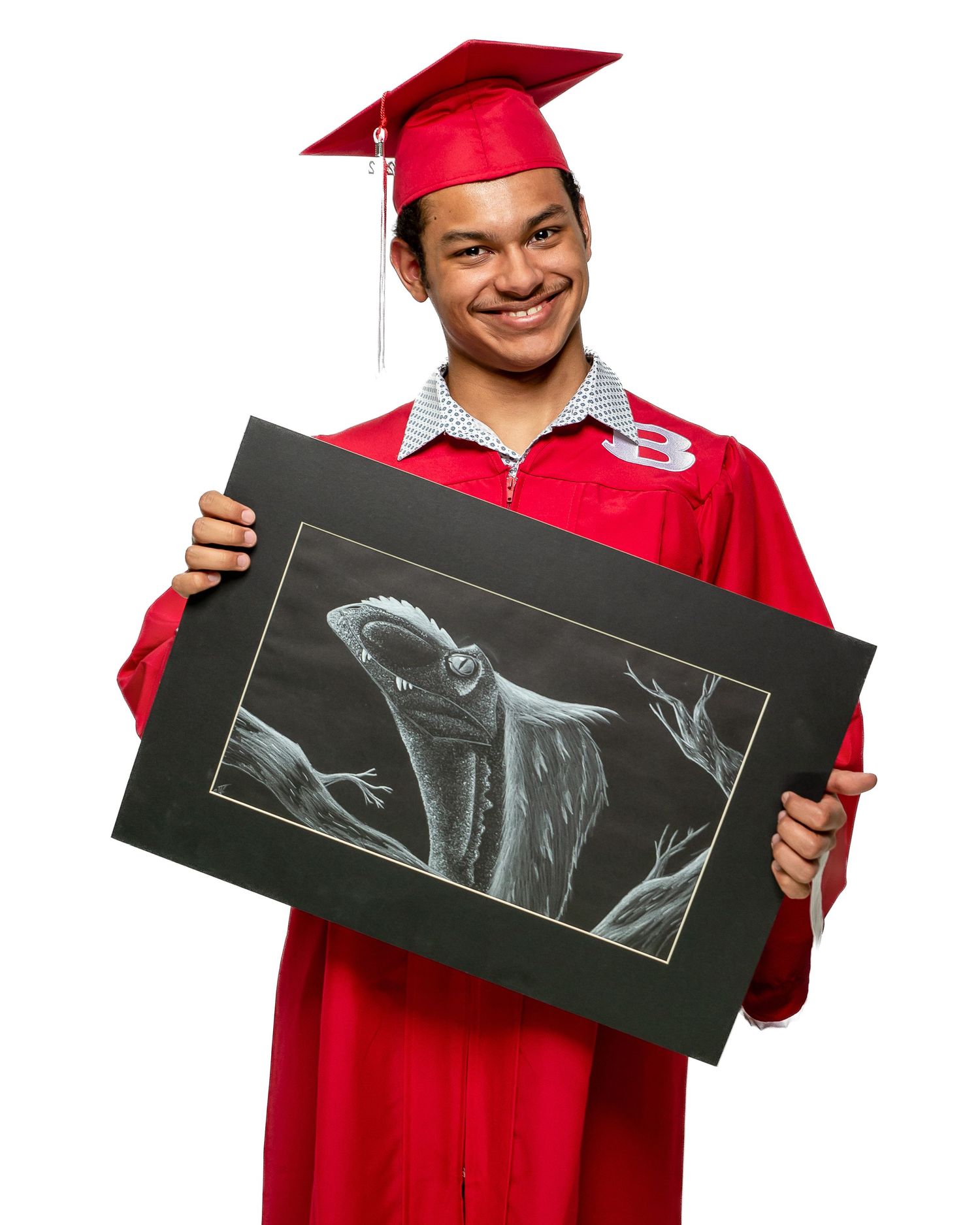 Smiling graduate in red cap and gown holds a detailed charcoal drawing of a lizard at graduation.