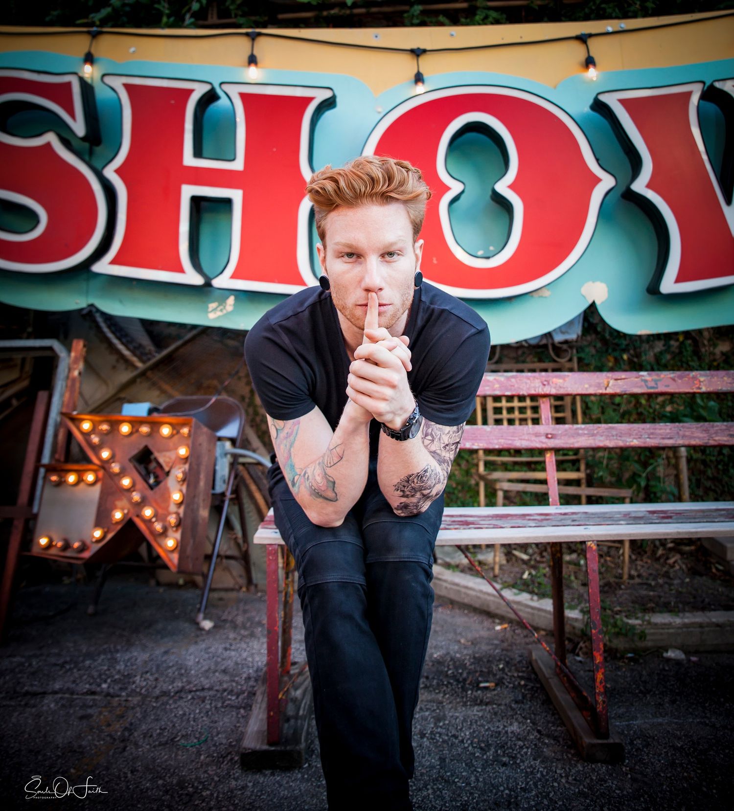 Tattooed man posing confidently in front of a colorful circus show sign outdoors.