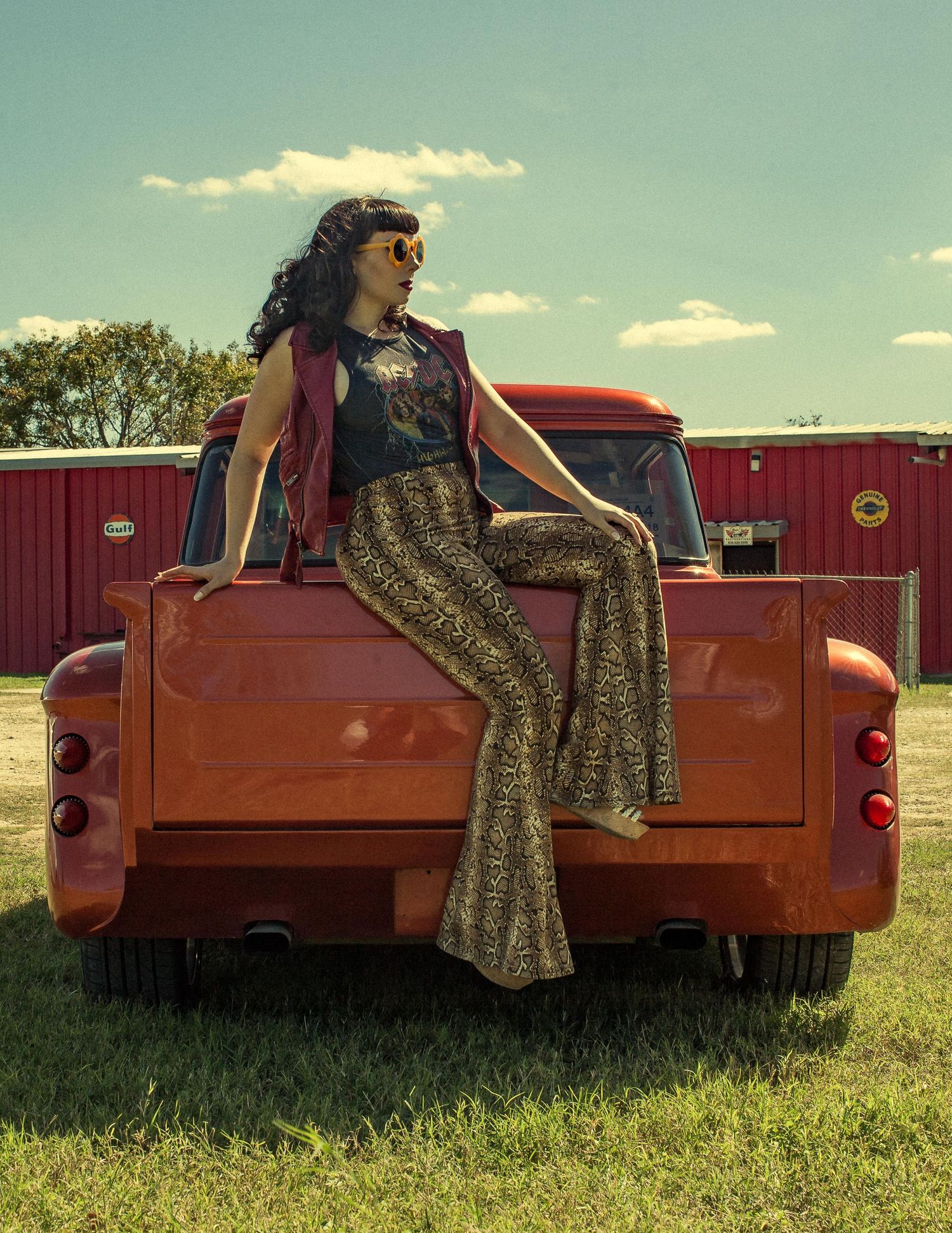 Woman in flare pants and sunglasses sitting on vintage orange pickup truck bed outdoors.