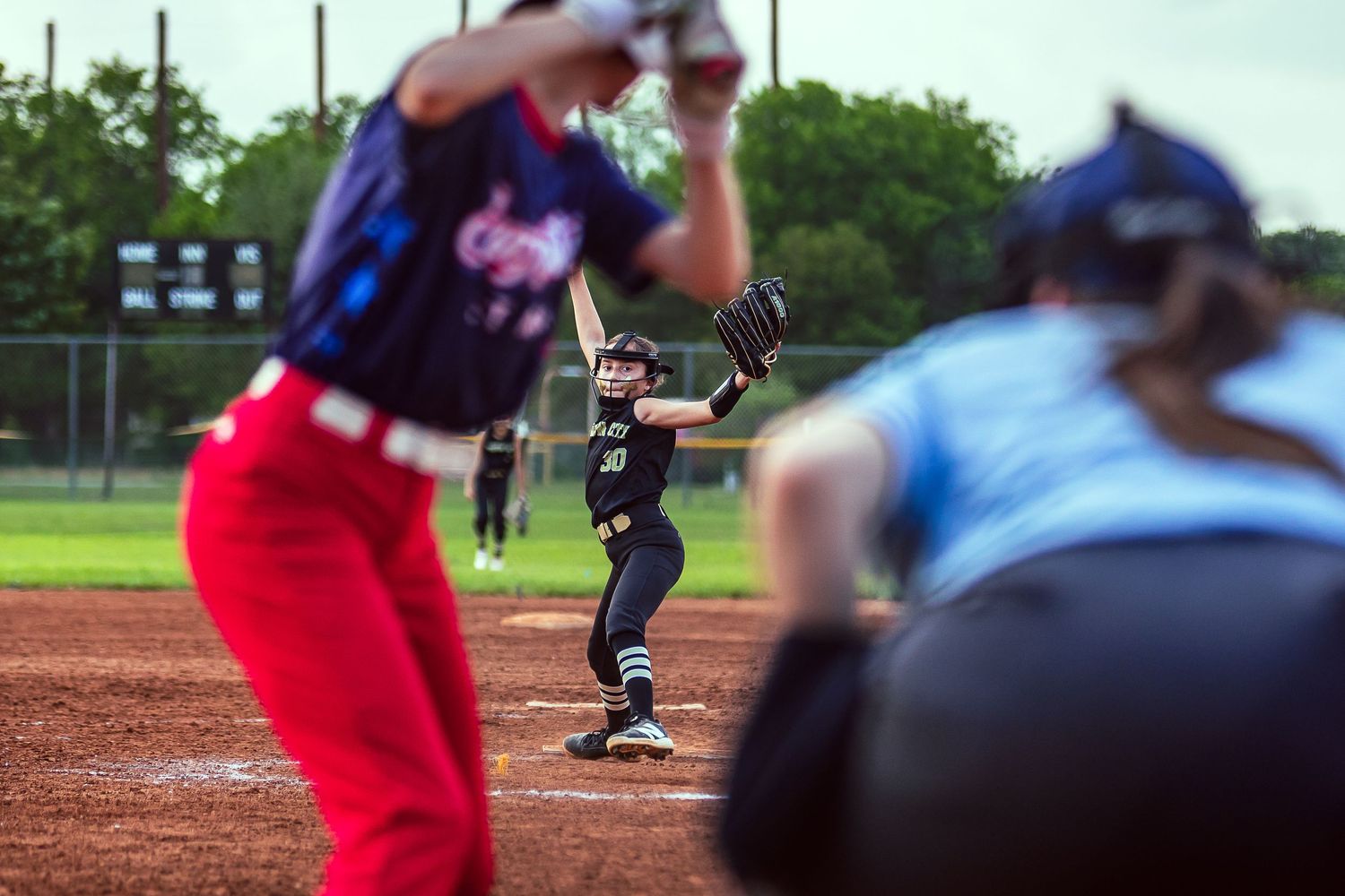 Female softball pitcher winds up to throw during a competitive game while batter and catcher await the pitch.