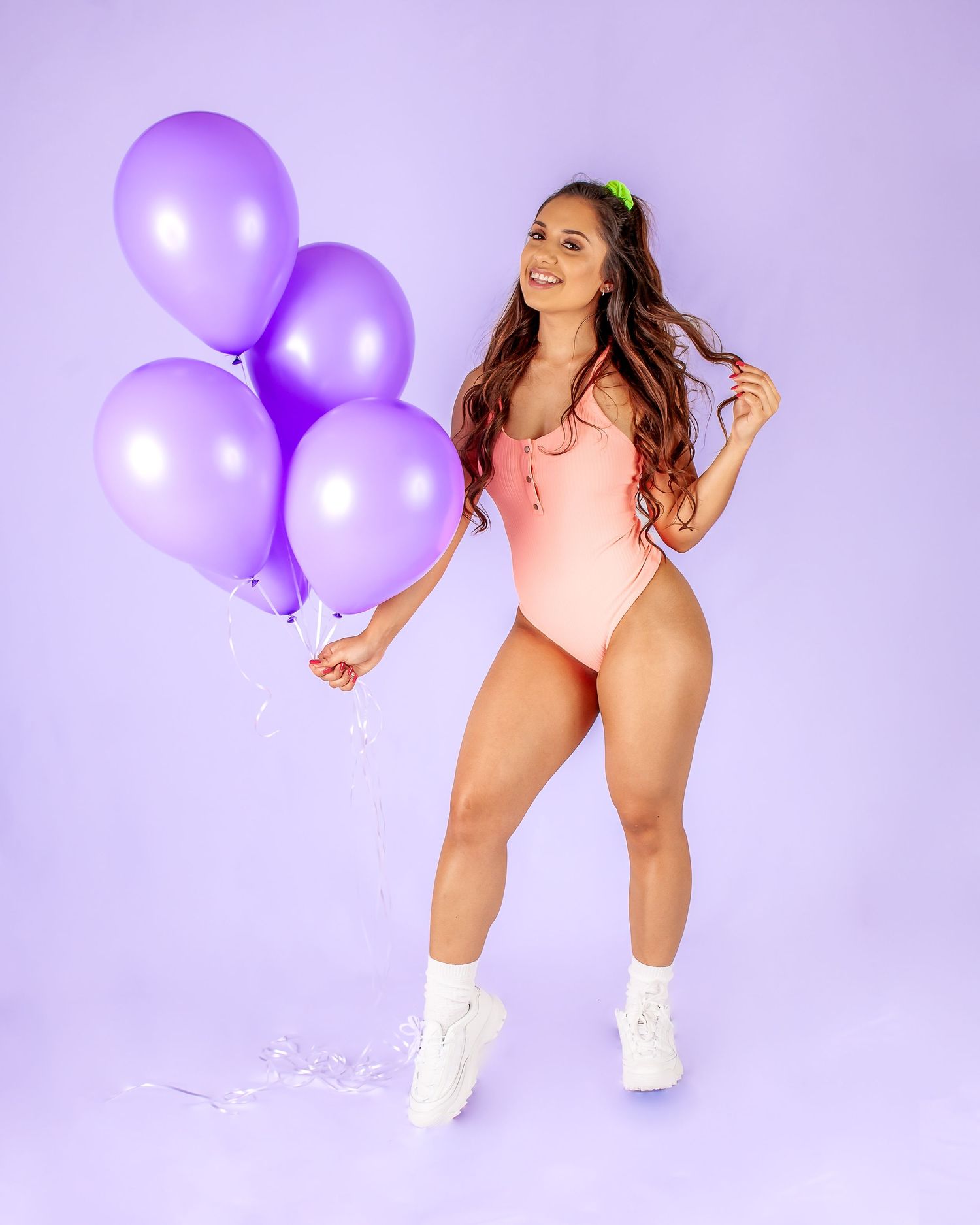 Woman in pink swimsuit holding purple balloons, wearing white sneakers, posing on purple background.