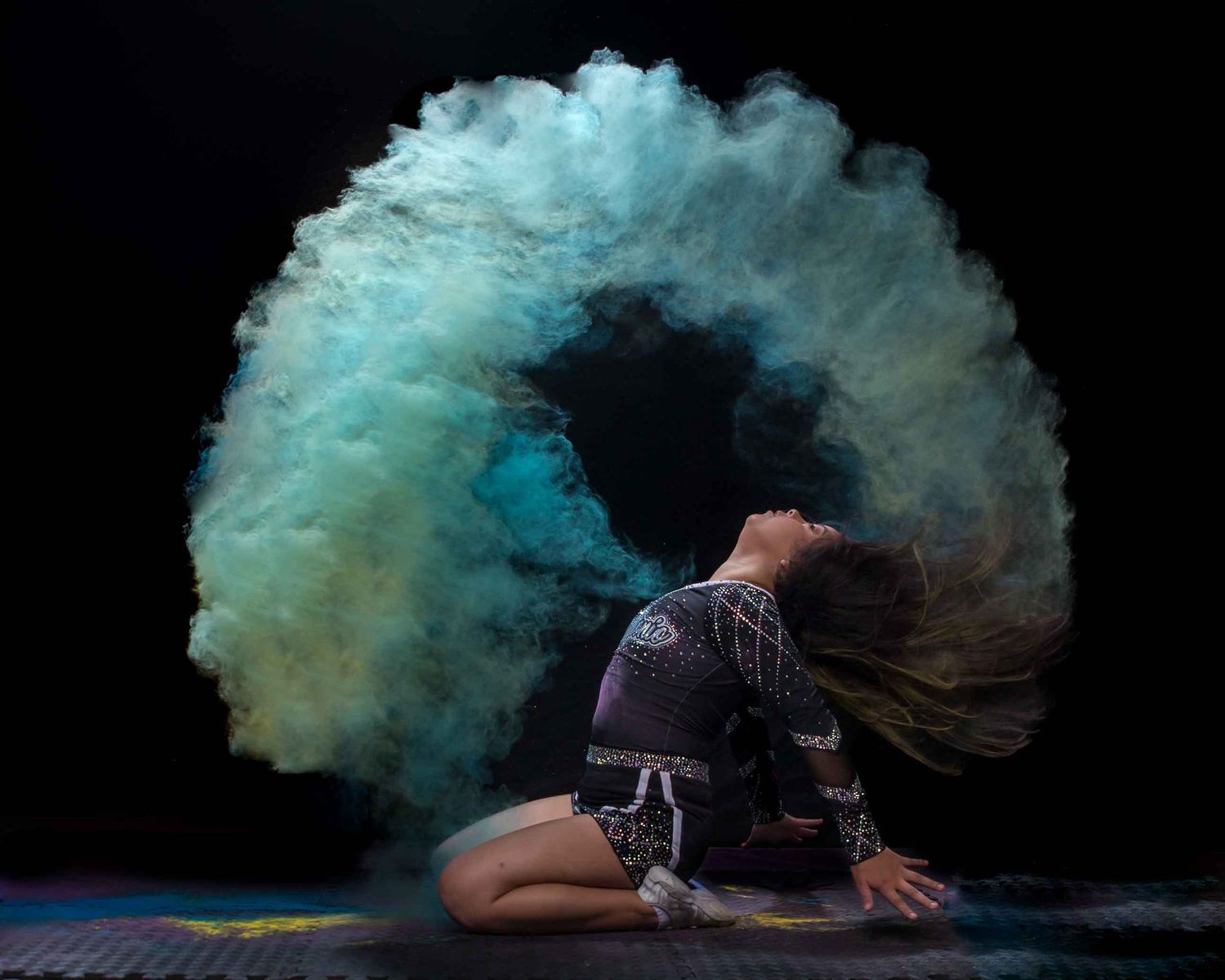 Gymnast kneeling on floor with dramatic teal and blue powder cloud explosion swirling around her against dark background.