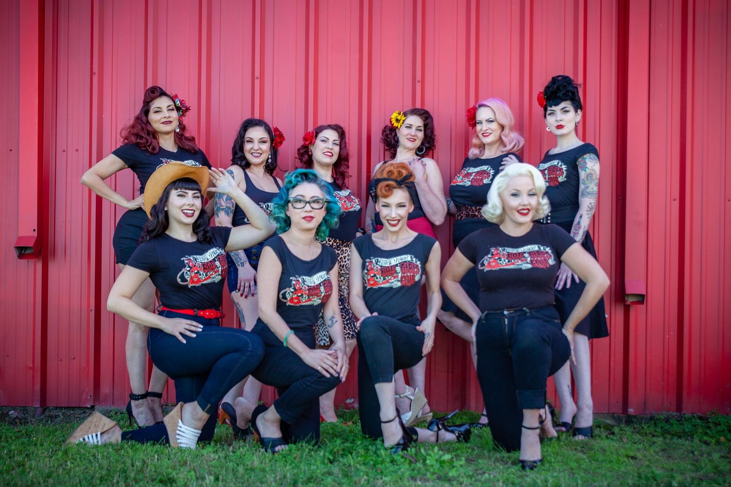 Group of women in matching black rockabilly outfits posing in front of a red barn wall outdoors.
