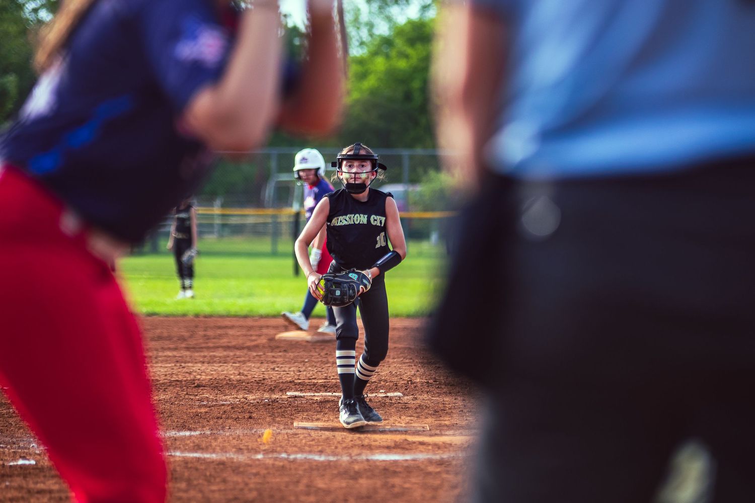 Softball player running bases during a competitive game on a dirt field with teammates and umpire visible.