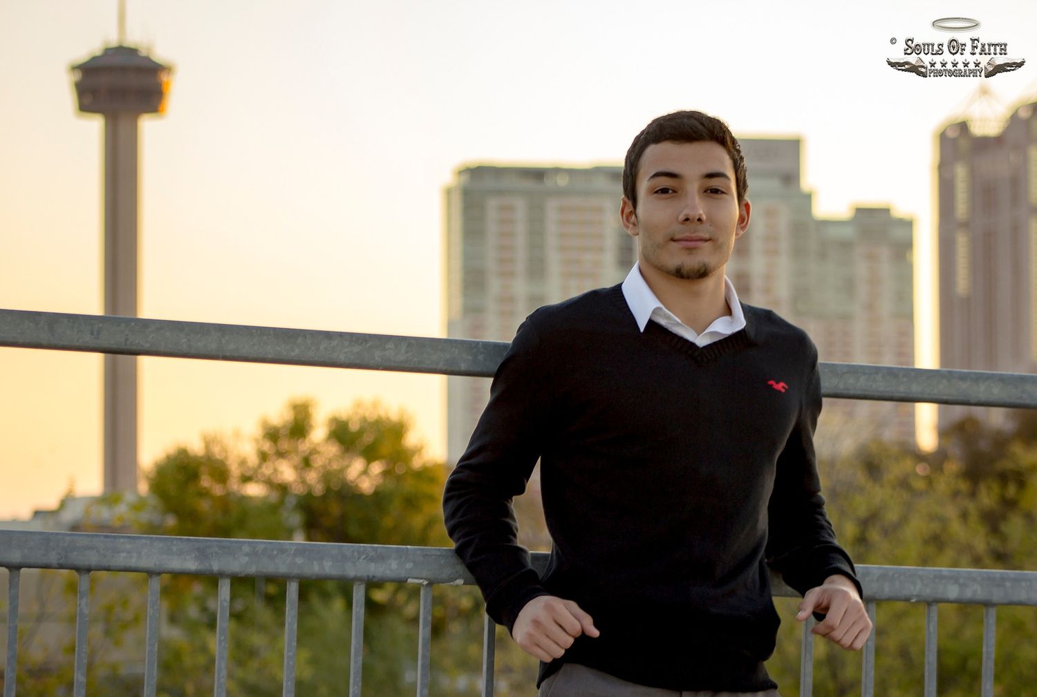 Young man in black sweater poses on a bridge with San Antonio Tower of the Americas skyline at golden hour.