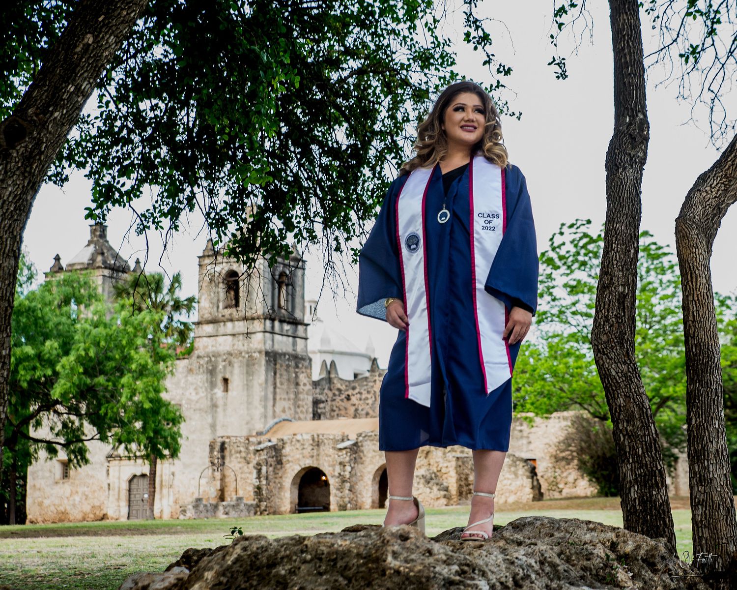 Woman in navy graduation gown and stole stands on rock near historic San Antonio mission on graduation day.