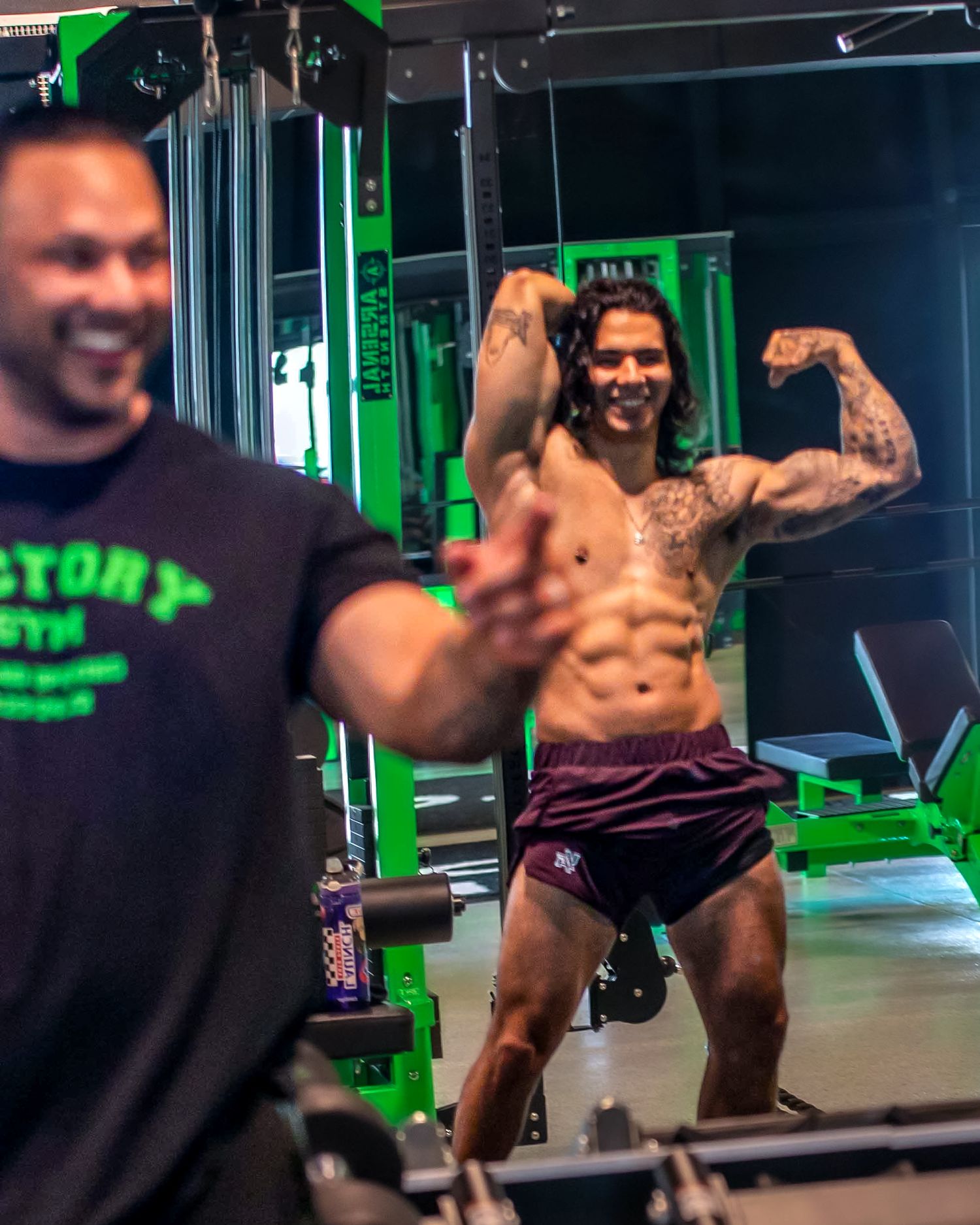 Tattooed muscular man flexing biceps shirtless in green-lit gym while another man in Victory Gym shirt smiles in foreground.