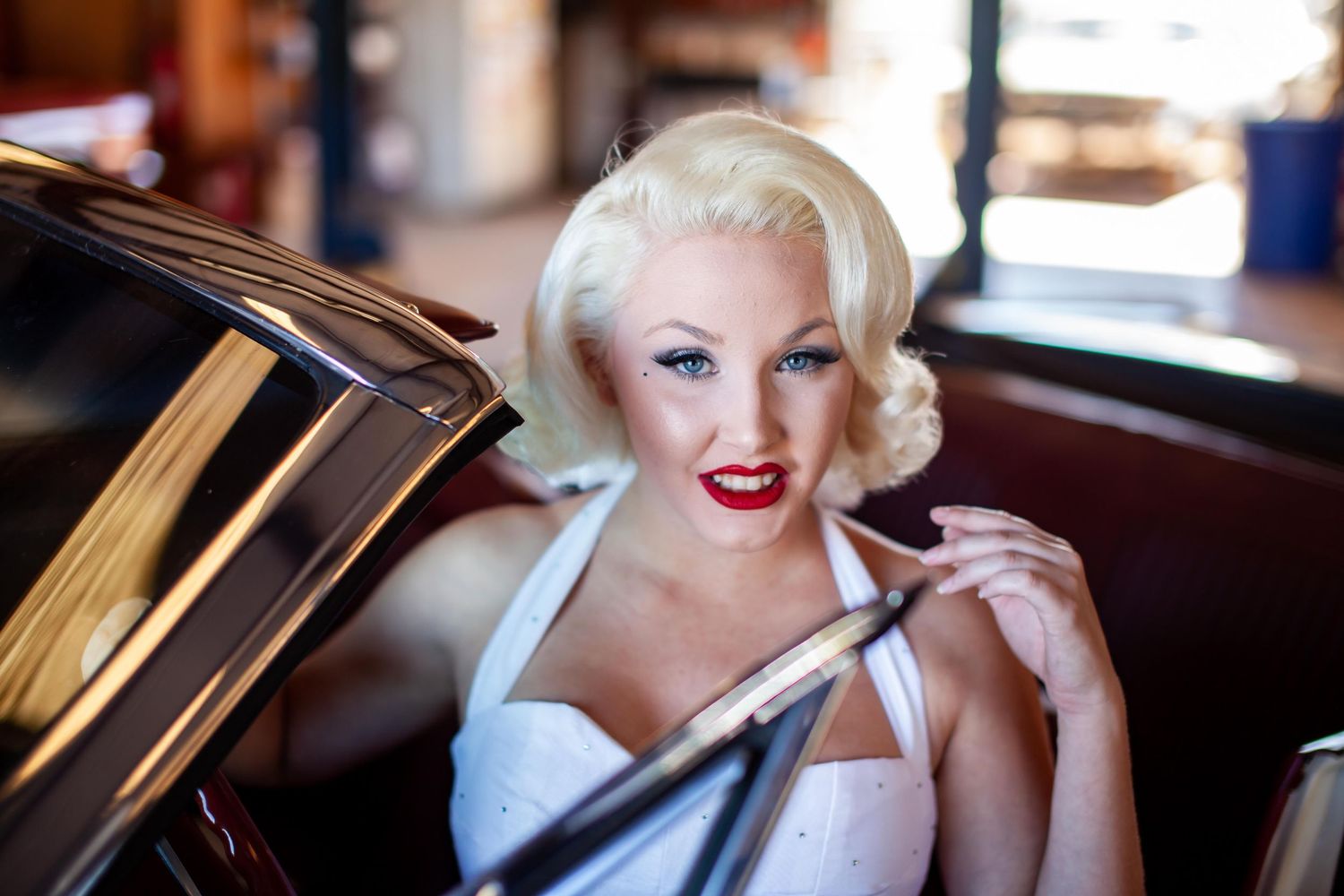 Blonde woman styled as Marilyn Monroe with red lips and white dress poses beside a vintage car.
