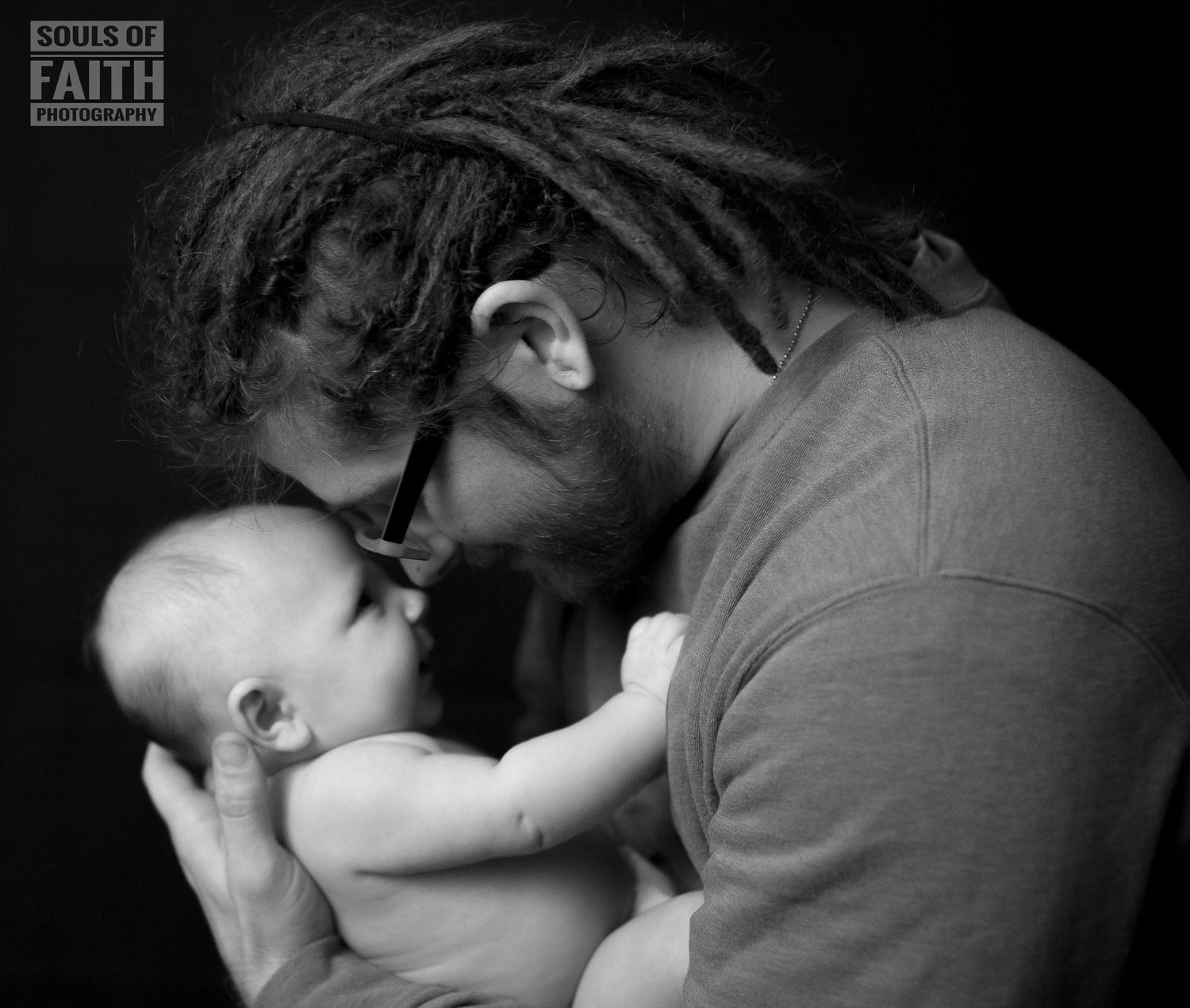 Black and white photo of a father with dreadlocks tenderly touching foreheads with his newborn baby.