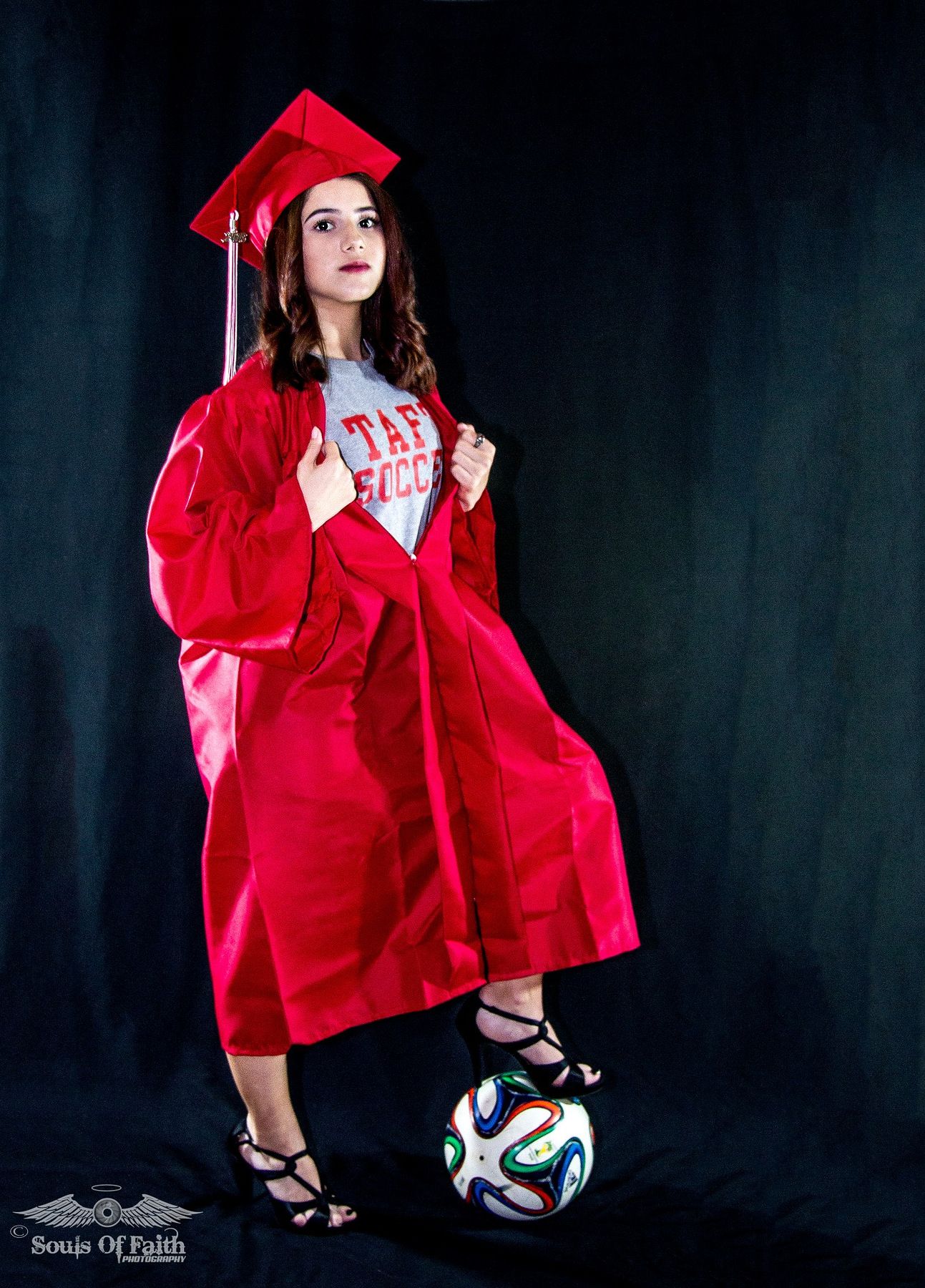Young woman in red graduation cap and gown posing with a soccer ball, wearing heels and a soccer shirt underneath.