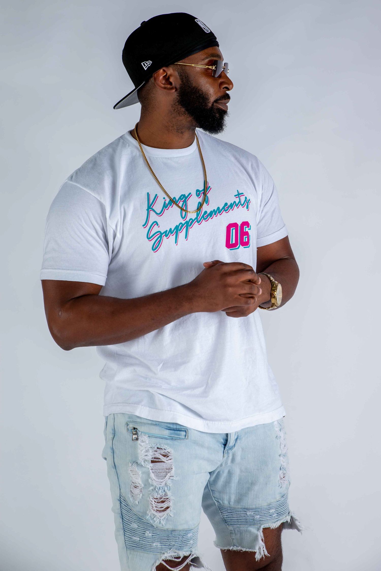 Man in white Kings Supplements 06 tee, ripped denim shorts, gold chain, snapback cap, posing confidently.
