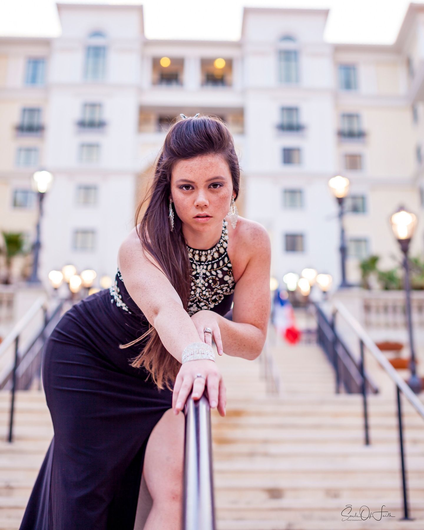 Young woman in black formal gown posing on steps near railing with elegant building backdrop at dusk.