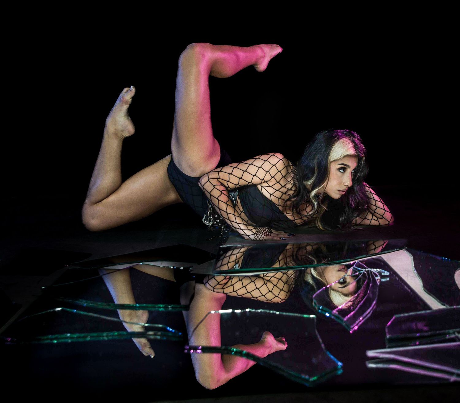 Woman in fishnet bodysuit poses dramatically on reflective surface with neon lighting in artistic photo.