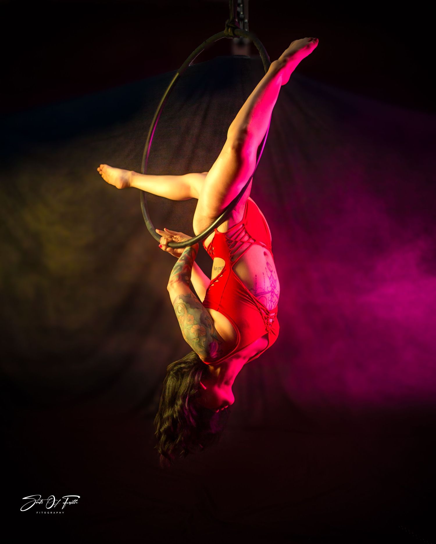Aerial hoop performer in red costume suspended upside down, illuminated by vibrant pink and yellow stage lighting.