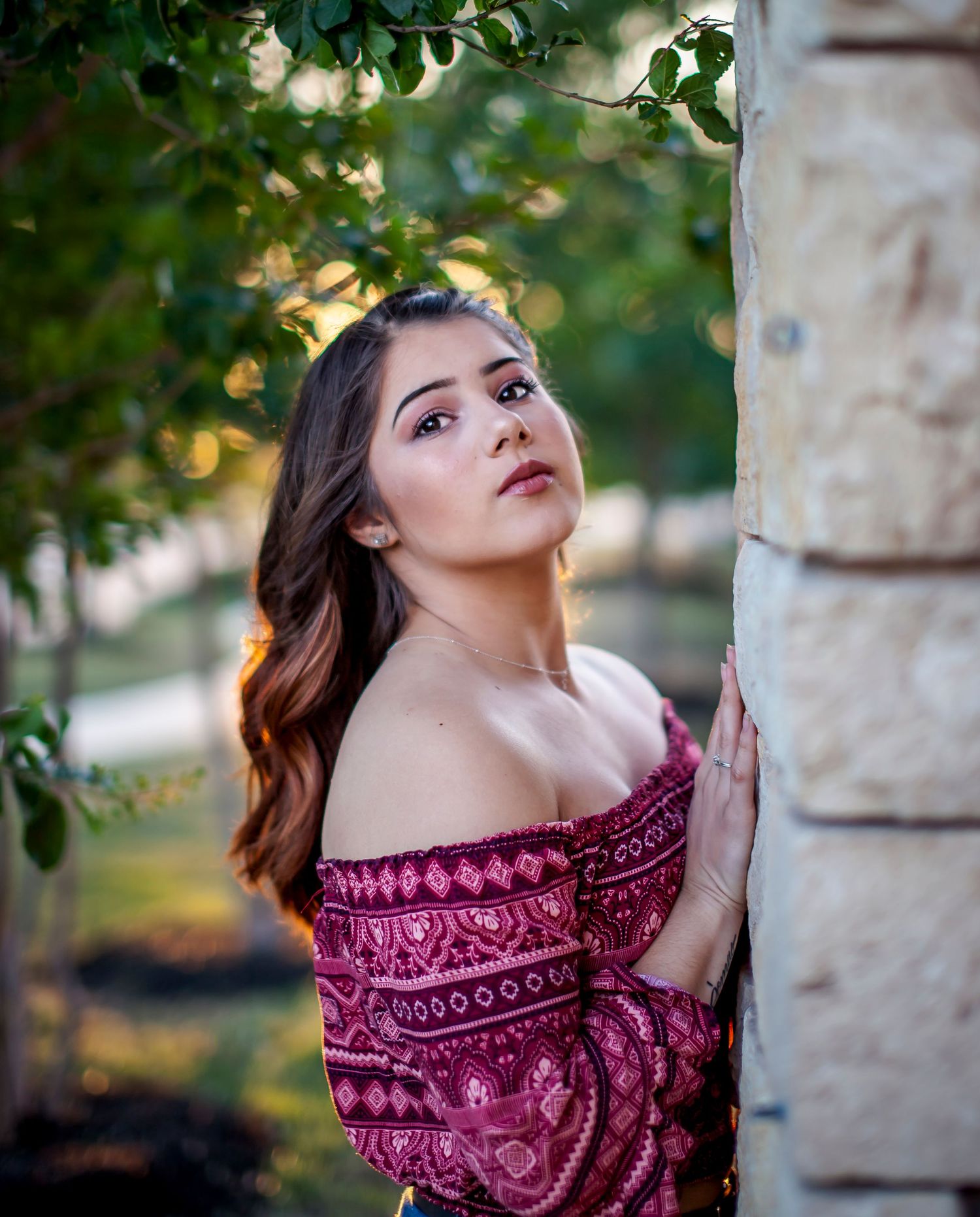 Young woman in red off-shoulder embroidered top leans against stone pillar surrounded by lush green trees. Golden hour senior photos.