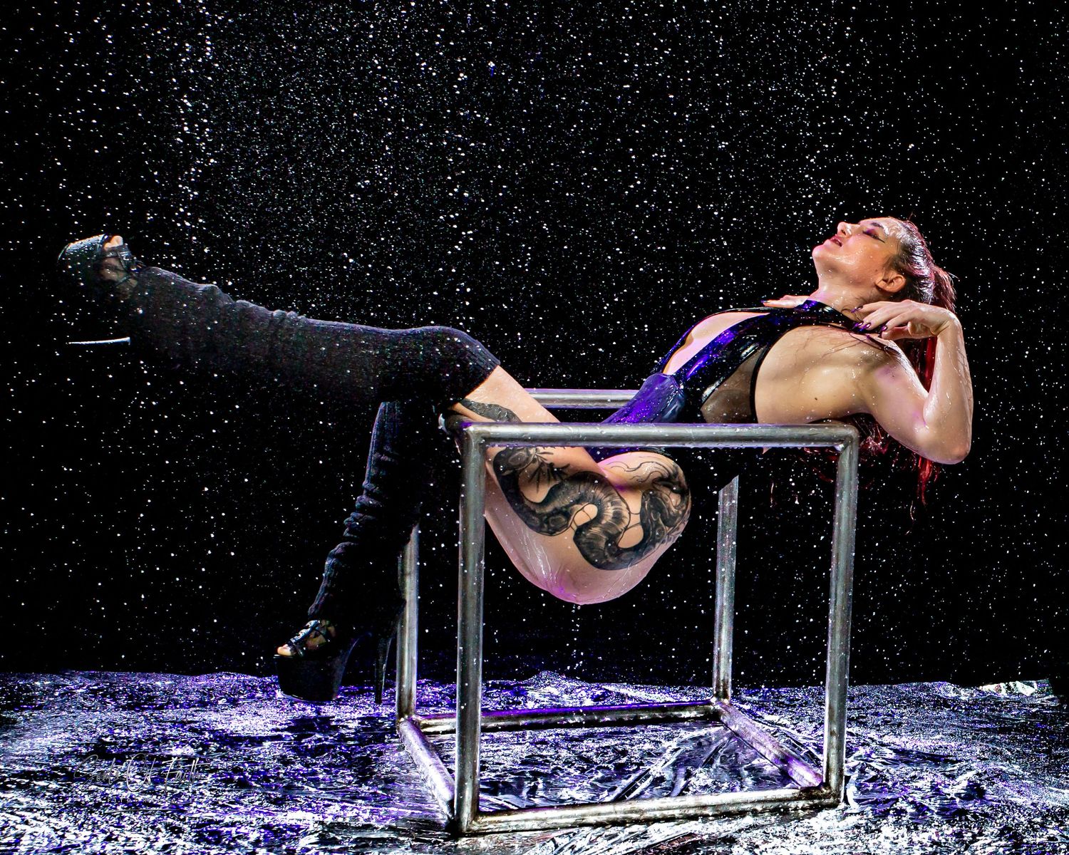 Tattooed performer reclines on metal frame prop under rain effects on dark stage in dramatic purple lighting.