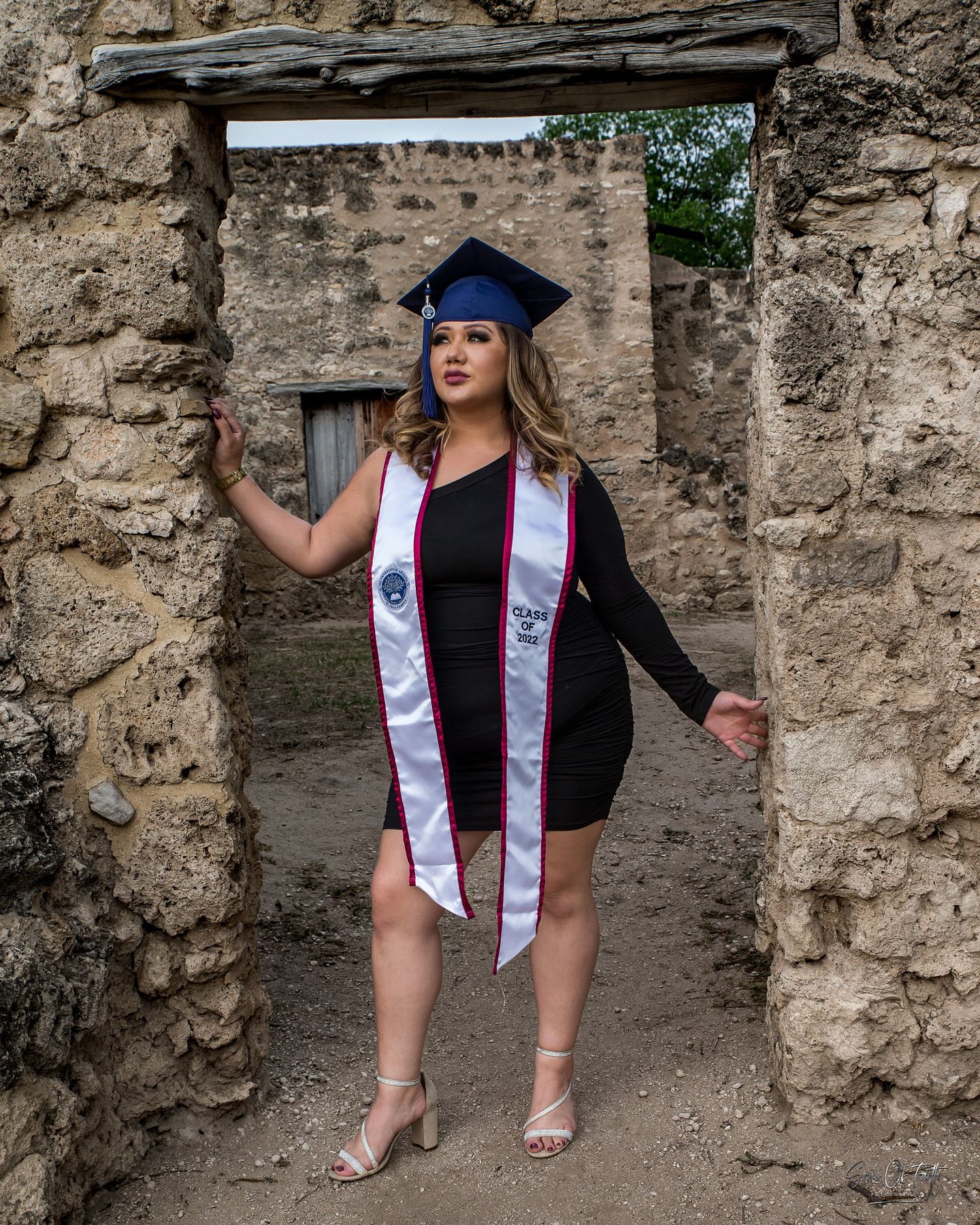 Young woman in graduation cap, stole, and black dress posing in stone archway for senior portrait.