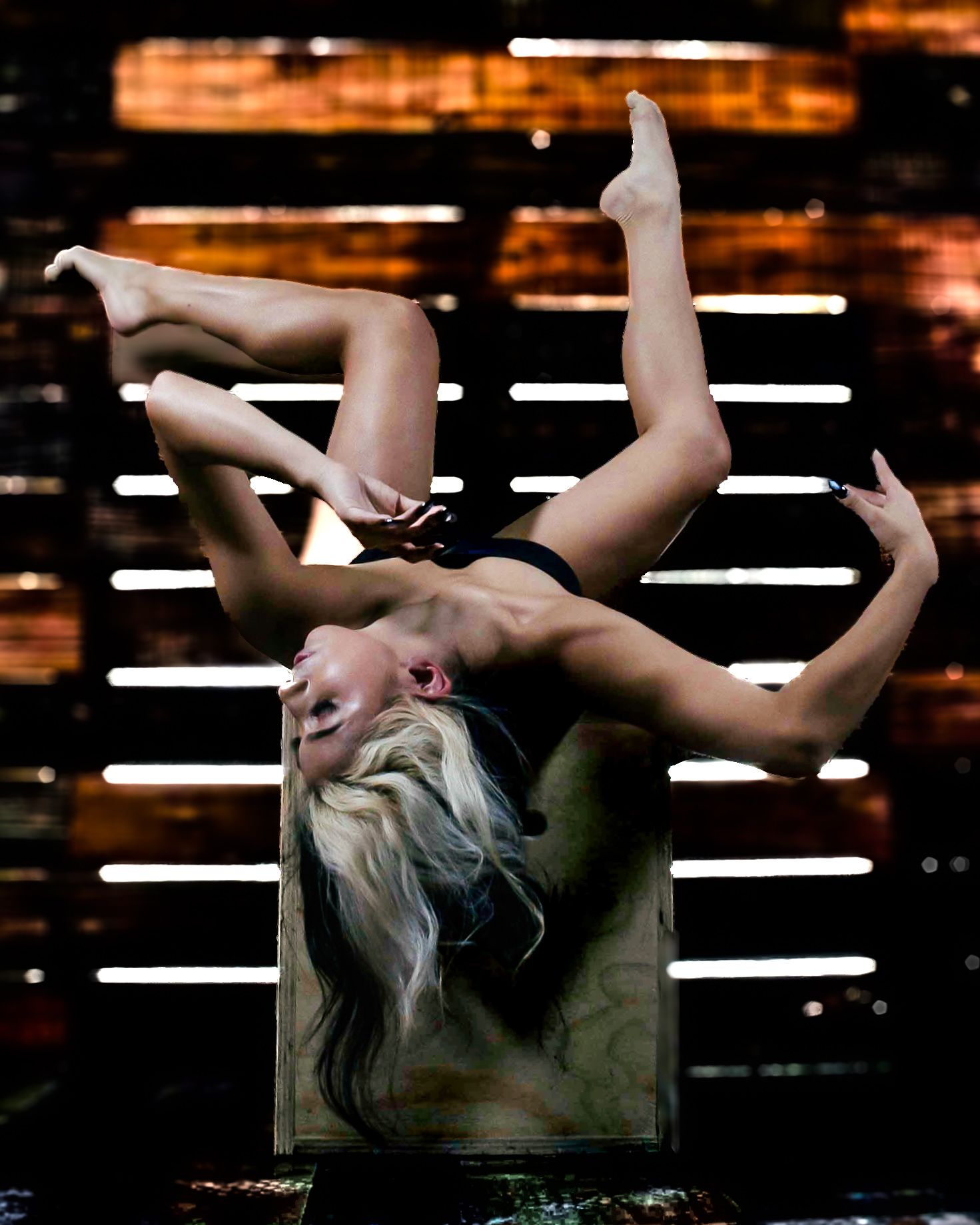Flexible blonde performer doing a backbend acrobatic pose on stage under dramatic lighting.