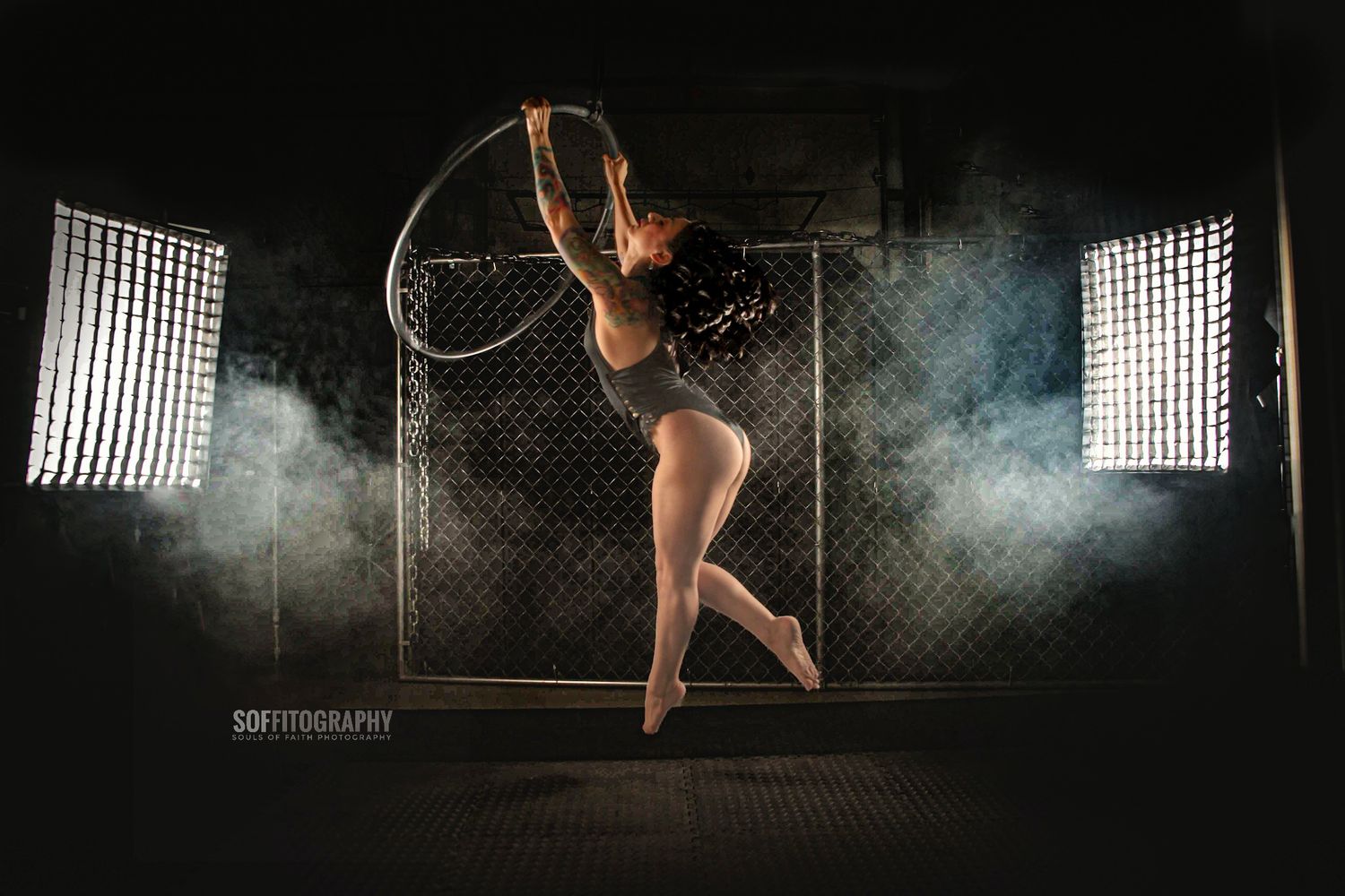 A dancer leaps gracefully holding a hula hoop in a dramatic studio shot with smoke and bright softbox lights.
