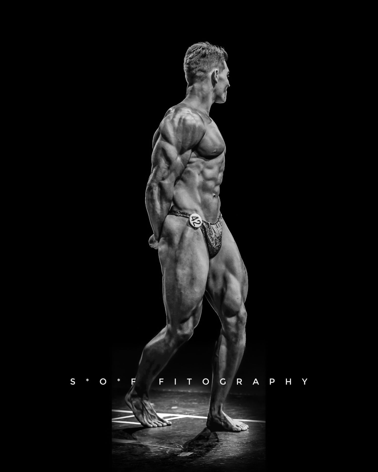 Black and white photo of a muscular male bodybuilder posing on stage, showcasing defined physique at a competition.