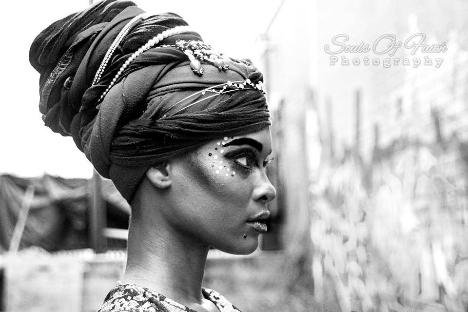 Black and white profile portrait of a woman wearing an elegant head wrap with decorative accessories and artistic face paint.