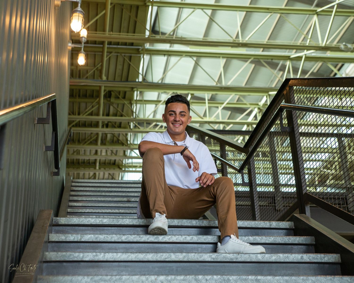 Young man in white shirt and tan pants sitting relaxed on modern industrial staircase with pendant lights.