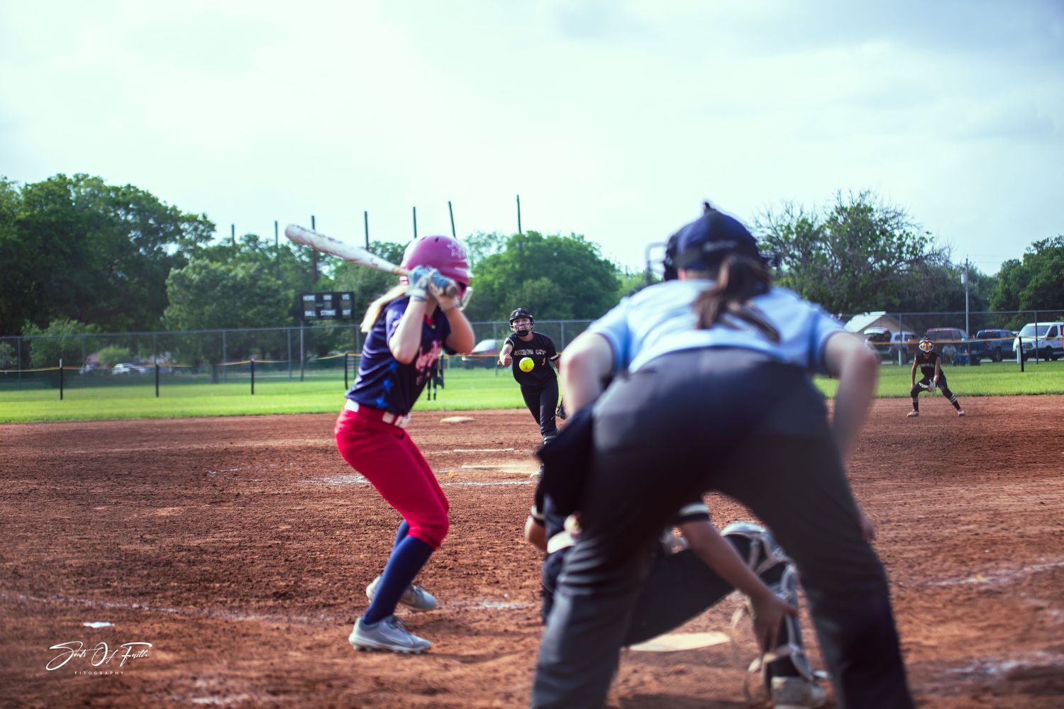 Female softball batter in red pants prepares to swing as catcher and umpire crouch behind home plate on dirt field.