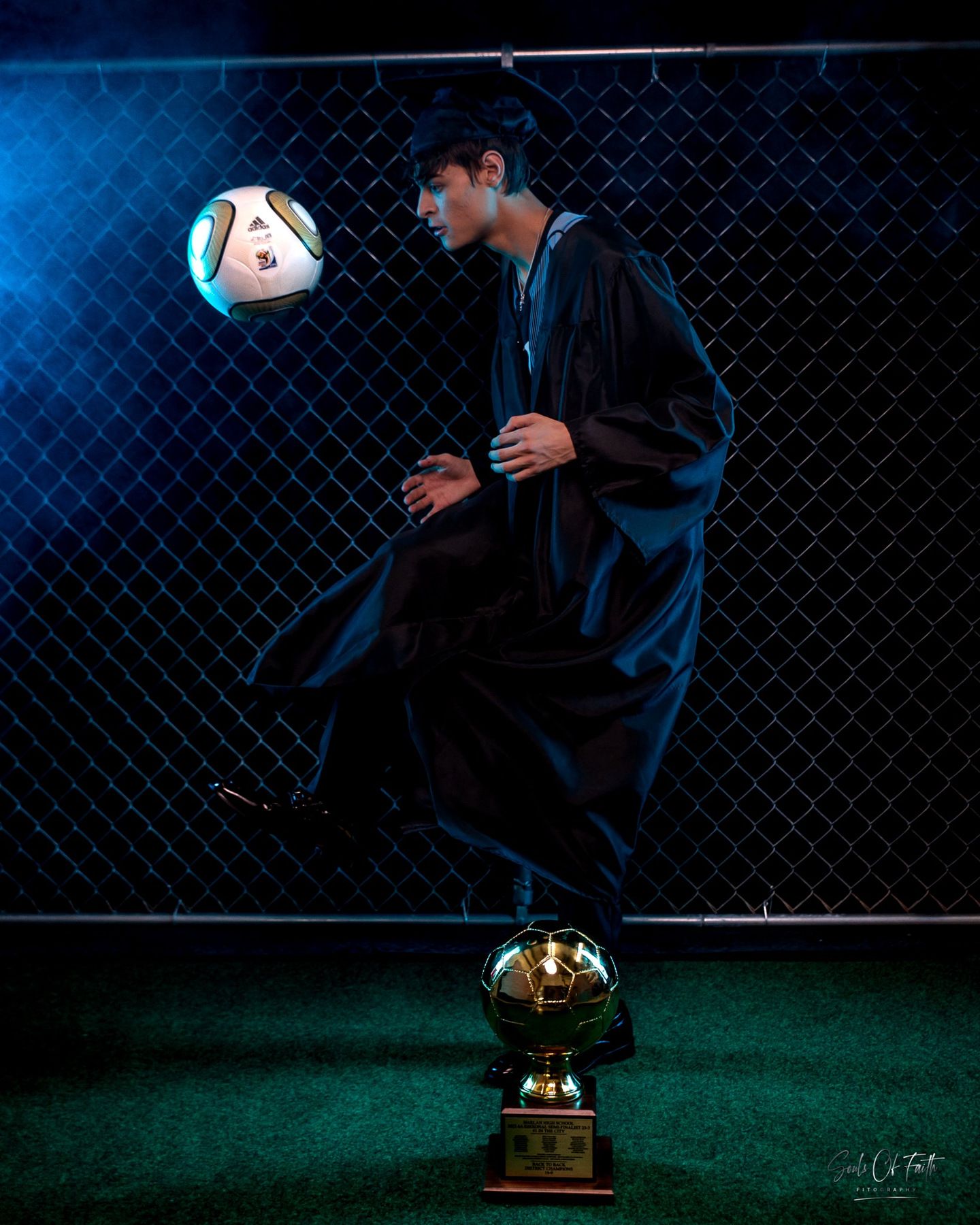 Graduate in cap and gown balancing soccer ball on knee beside golden trophy by chain-link fence.