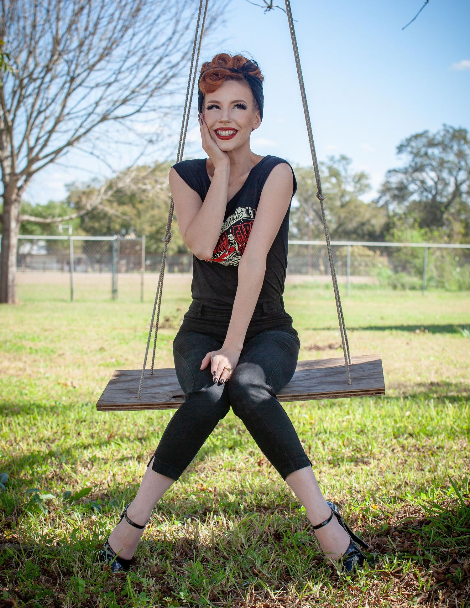 Woman with red updo sits on wooden swing outdoors, wearing black outfit with decorative top, smiling confidently.