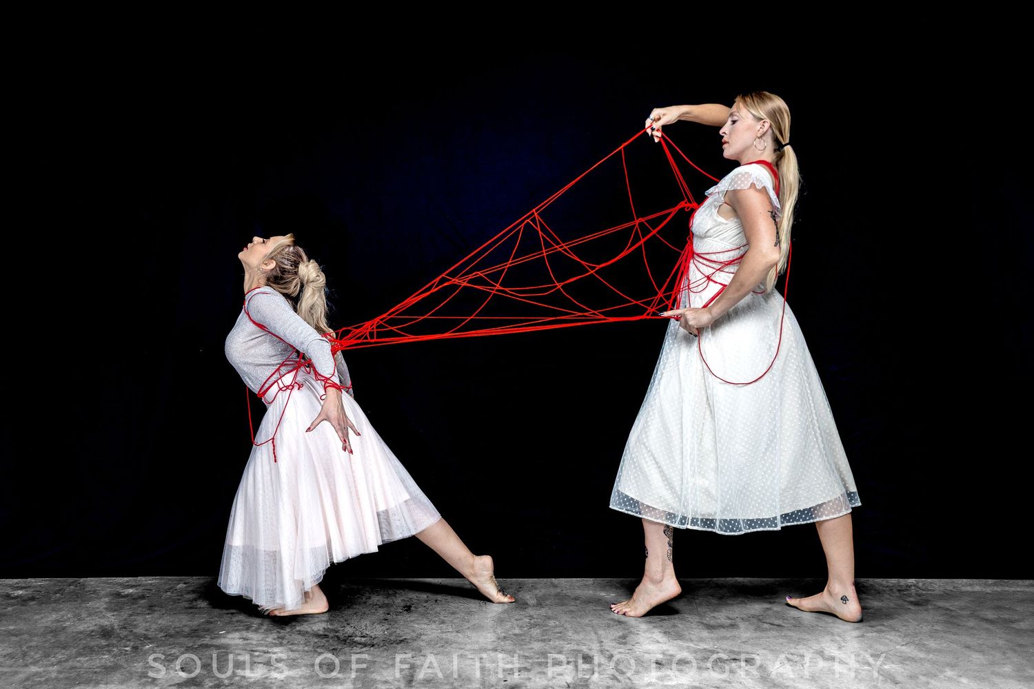 Two dancers in white dresses connected by red string in dramatic artistic performance photography.