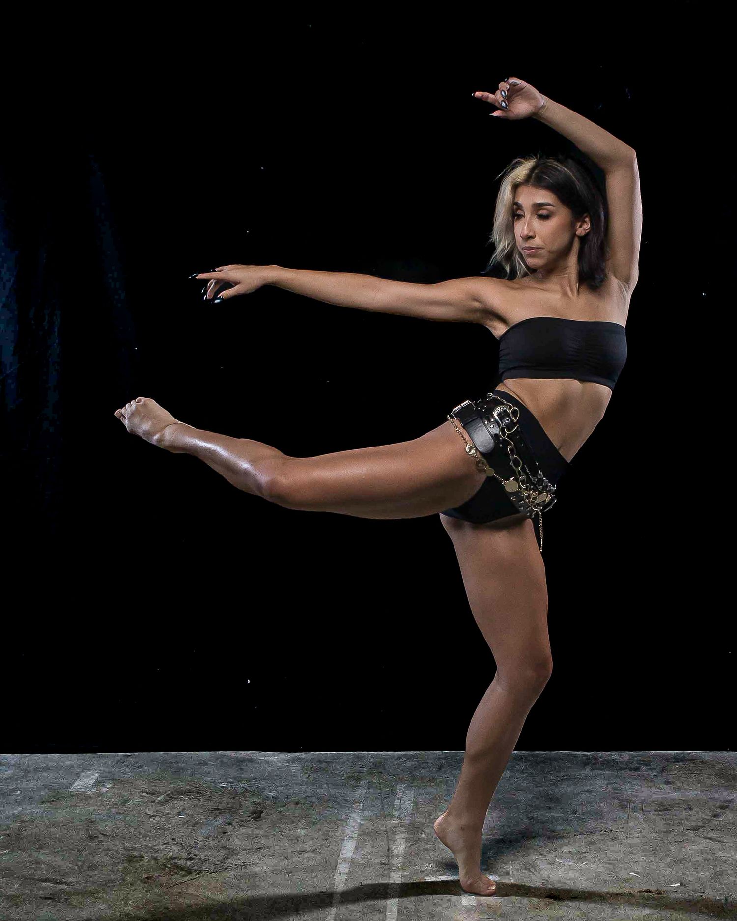 A dancer performs a high kick in a black crop top and shorts against a dark studio background.