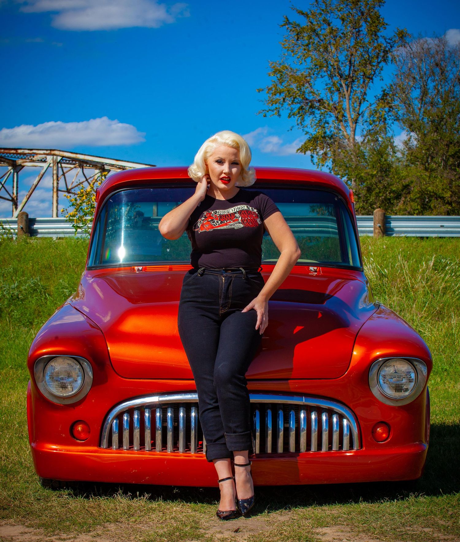 Blonde woman in retro pin-up style poses leaning on a classic red custom car outdoors on a sunny day.