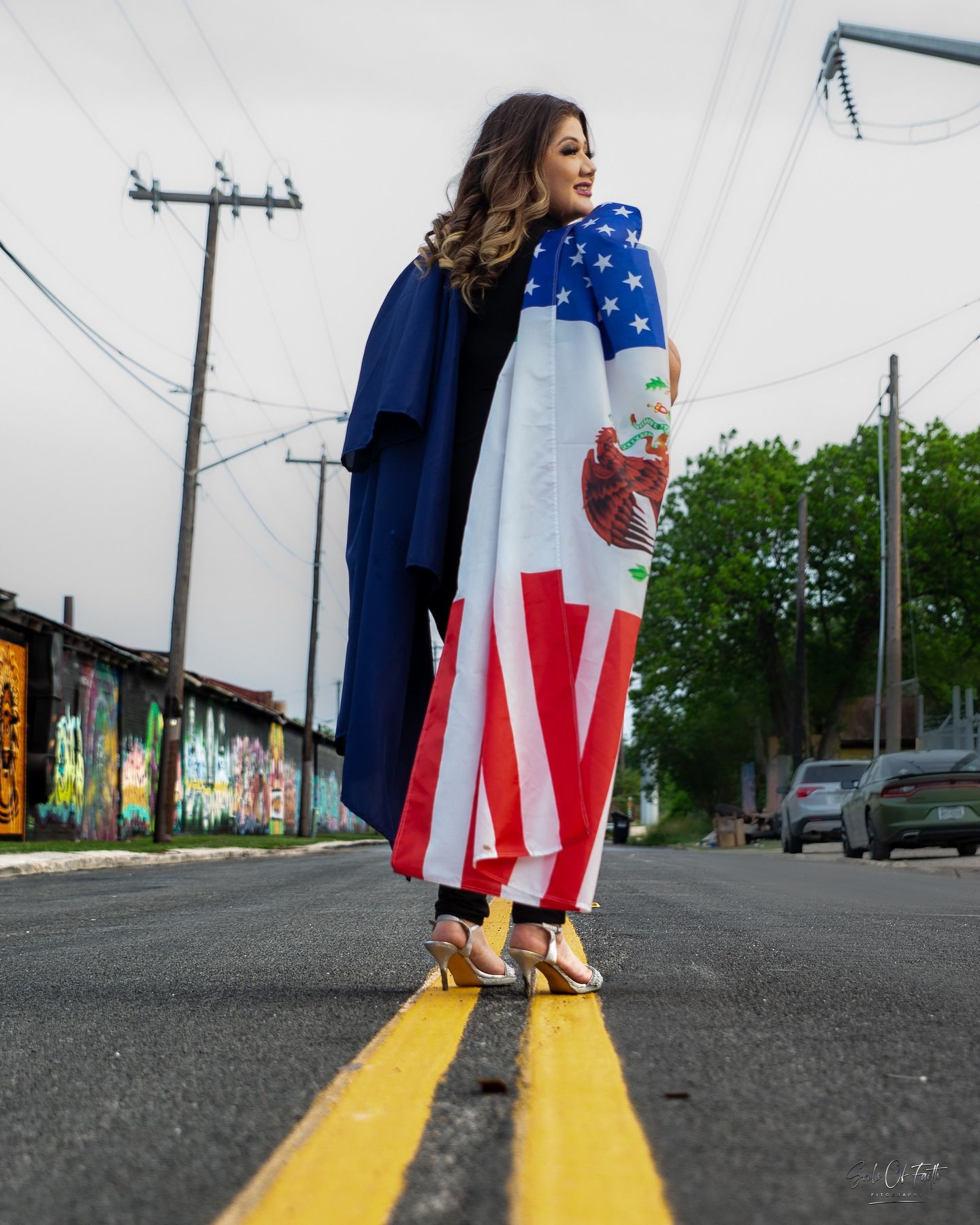 Woman in graduation gown draped with American and Mexican flags standing on a street road.