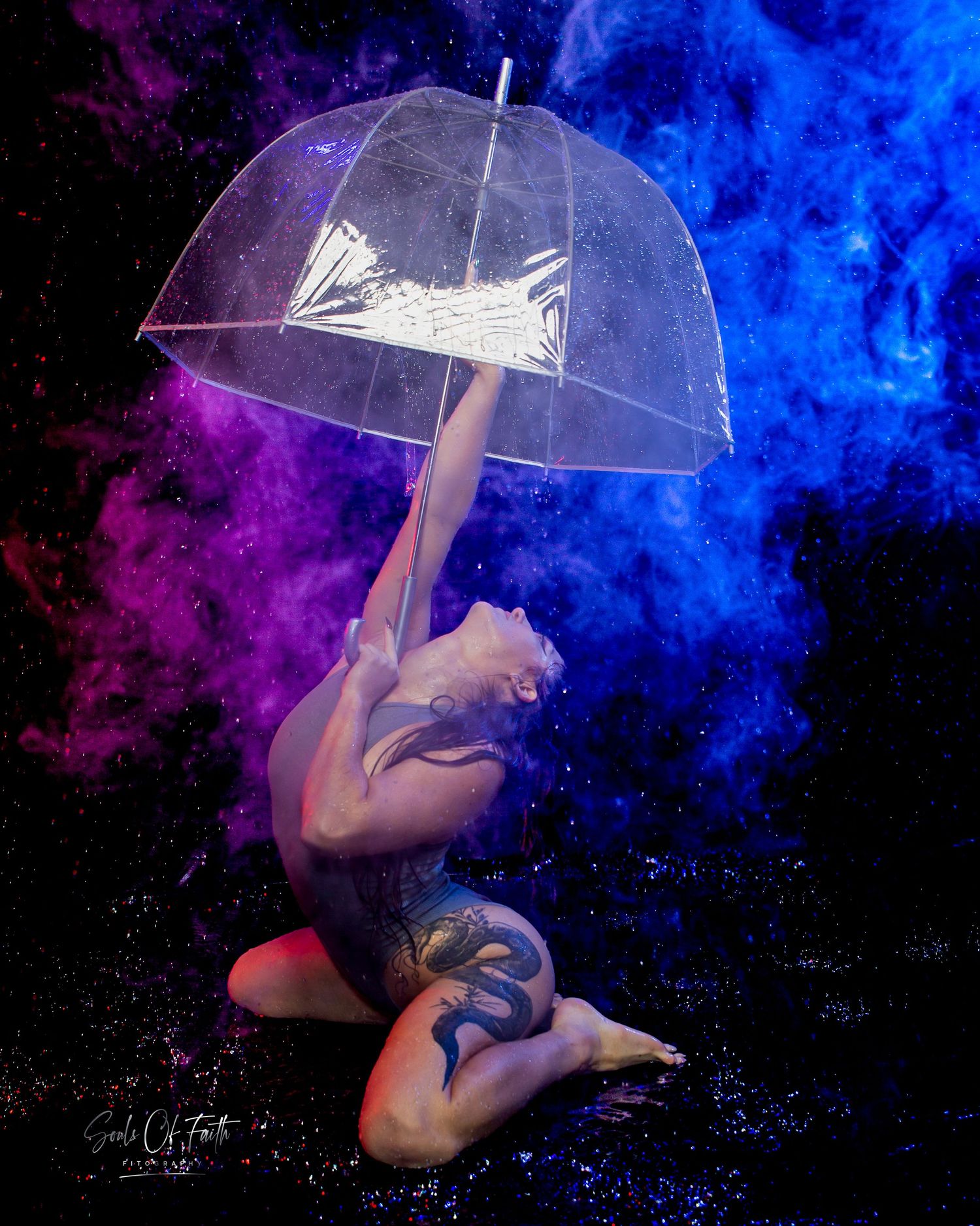 Tattooed performer kneeling dramatically holding clear umbrella amid purple and blue smoke clouds on dark background.