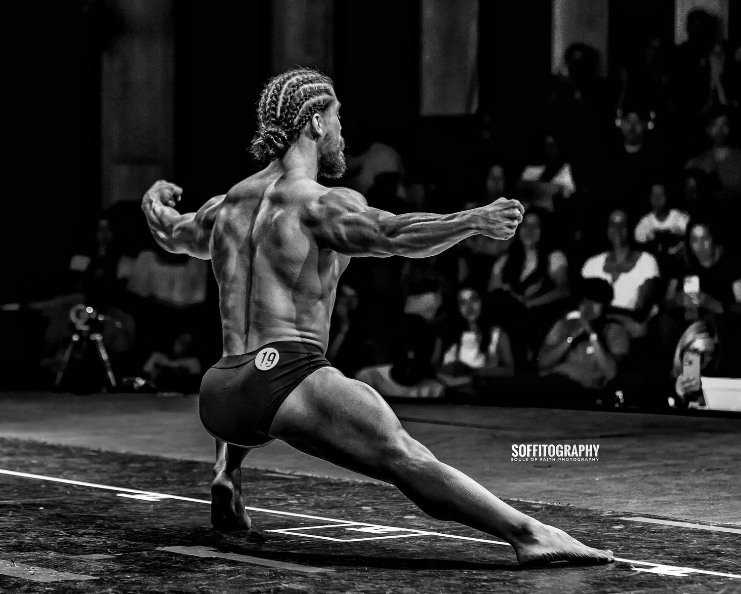 Black and white photo of a muscular male bodybuilder posing on stage in a lunging stance at a competition.