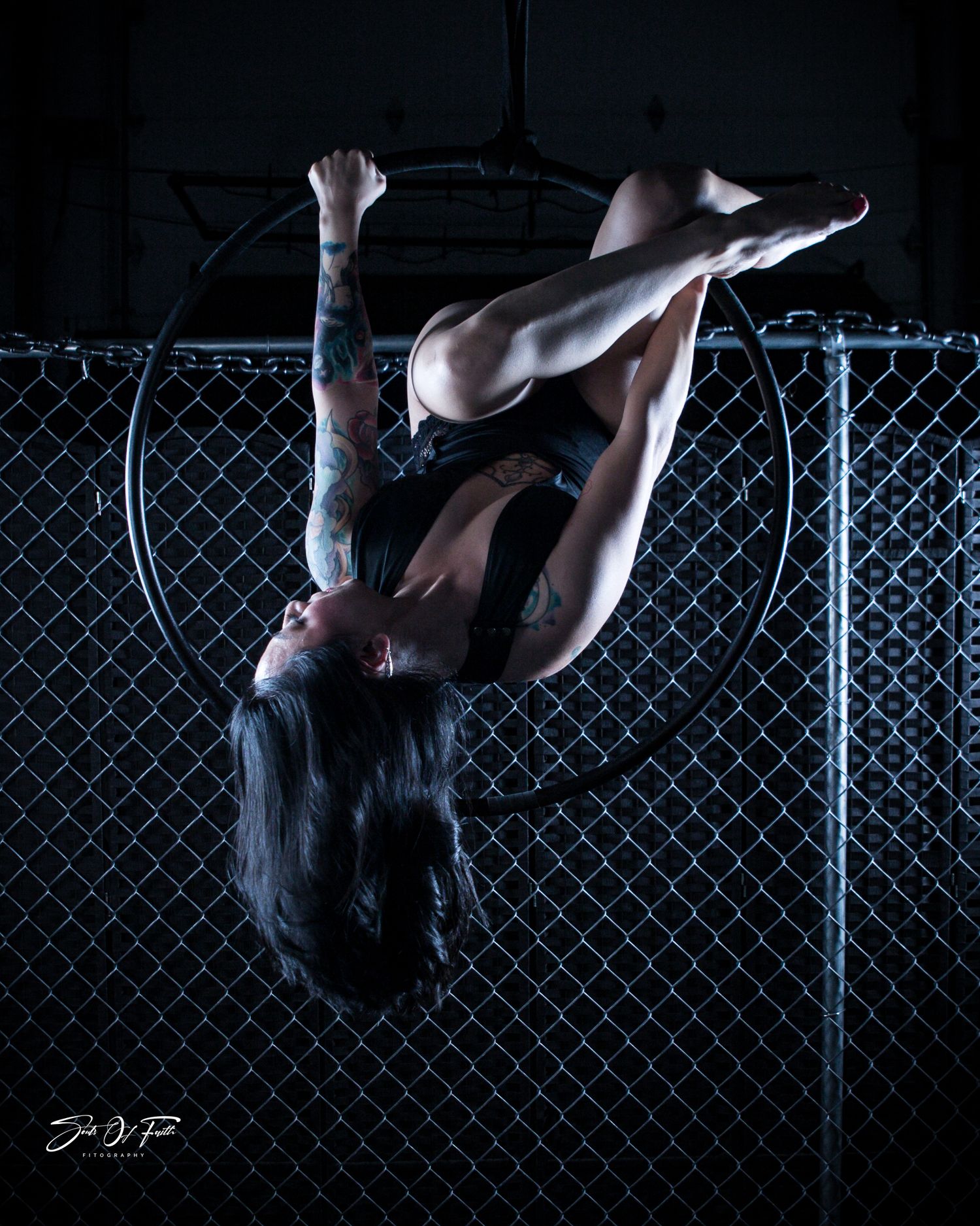Tattooed aerial hoop performer executing an inverted split pose on a lyra against a chain-link fence backdrop.