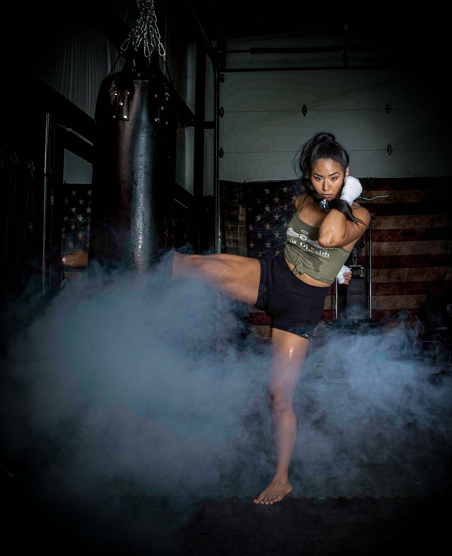 Female MMA fighter executing a high kick in a smoky gym with a punching bag and American flag backdrop.