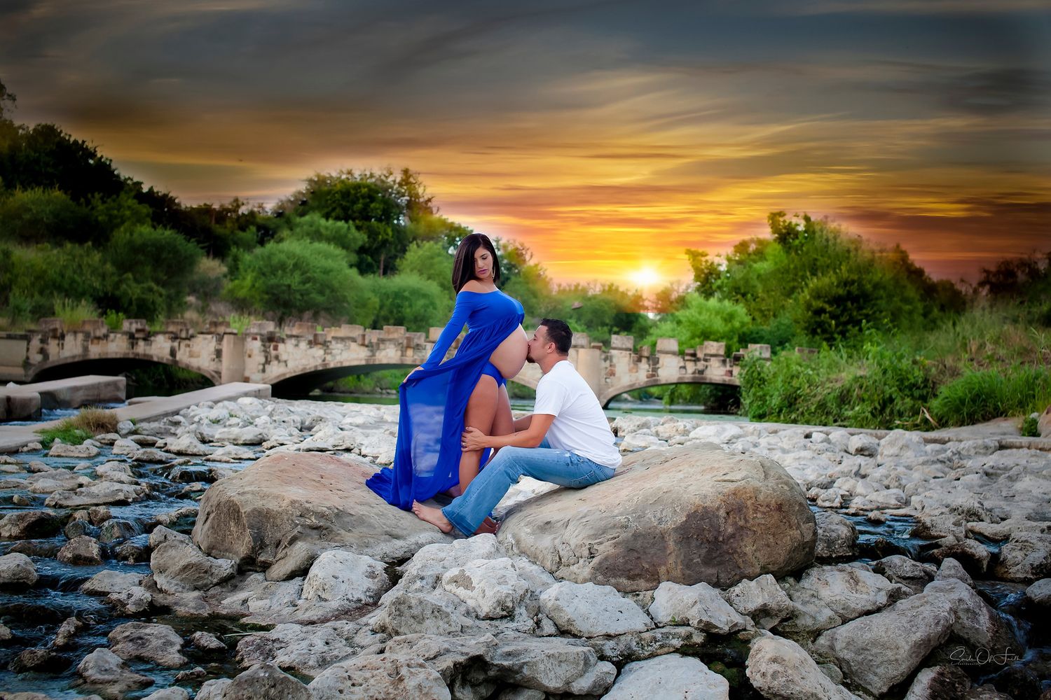 Couple sharing a romantic moment on rocks by a stream at sunset, woman in blue dress, vibrant sky backdrop.