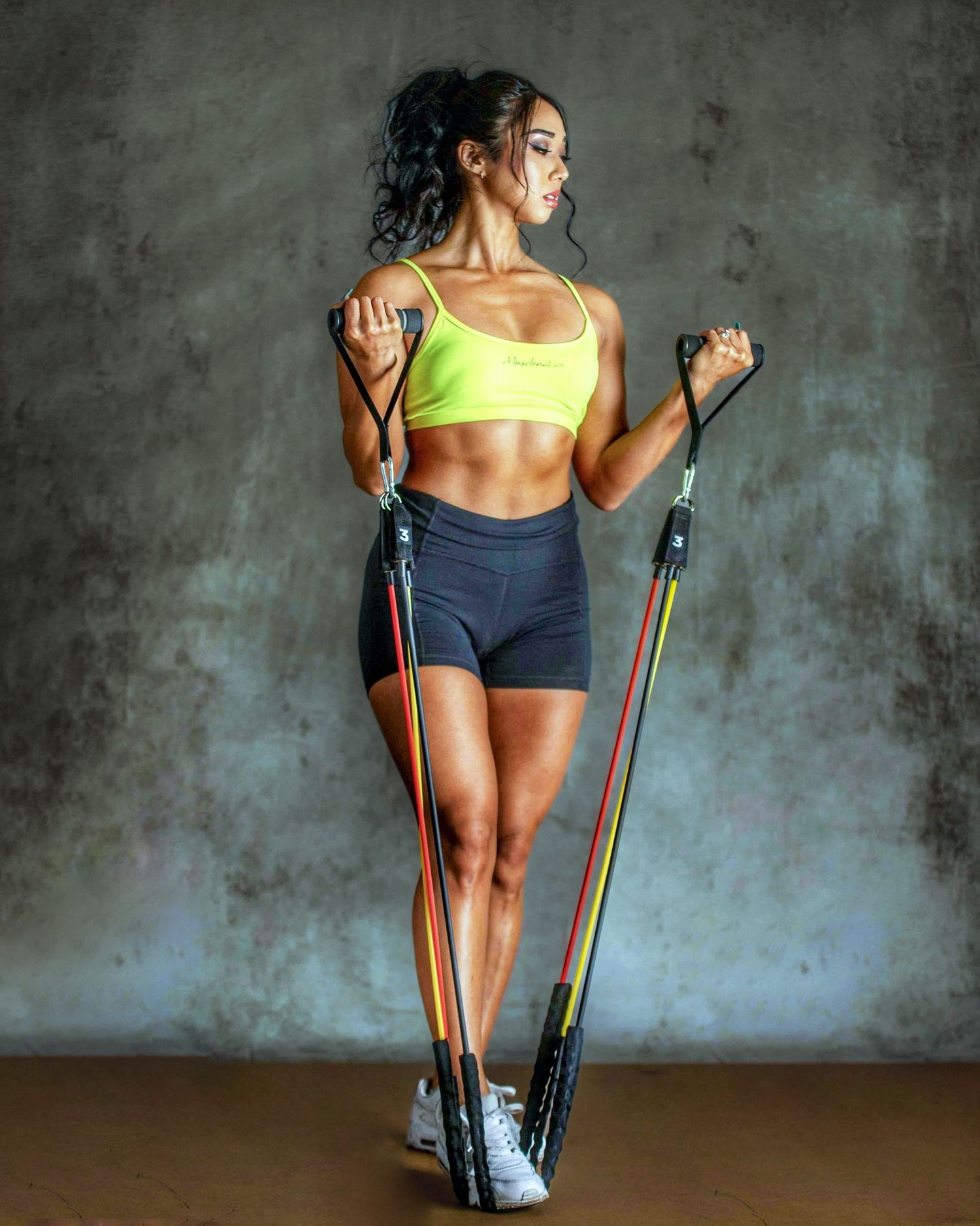 Fit woman in yellow sports bra and shorts using resistance bands for a full-body workout against a gray wall.