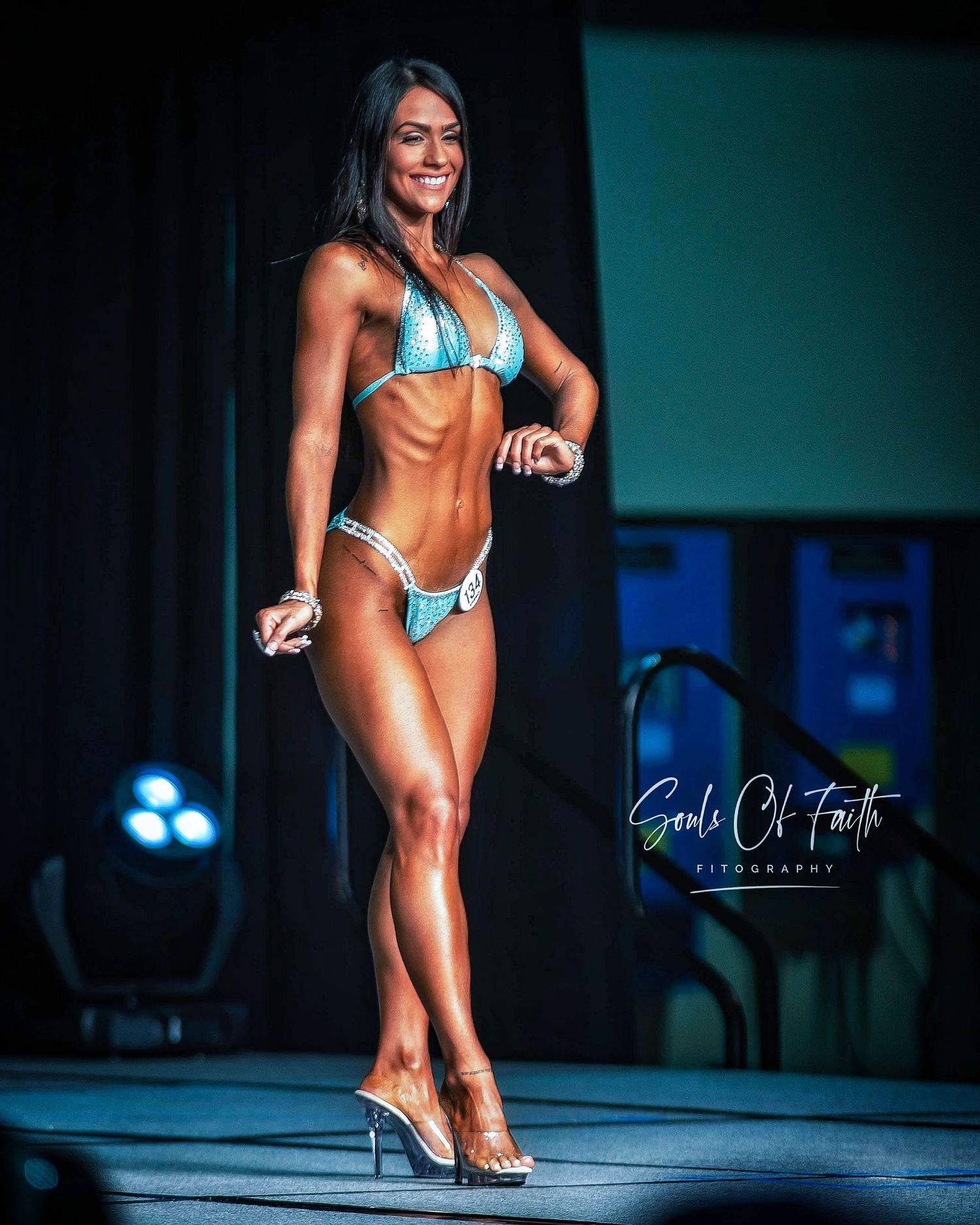 Female fitness competitor poses on stage in a sparkling teal bikini and heels under bright lights at a bodybuilding competition.