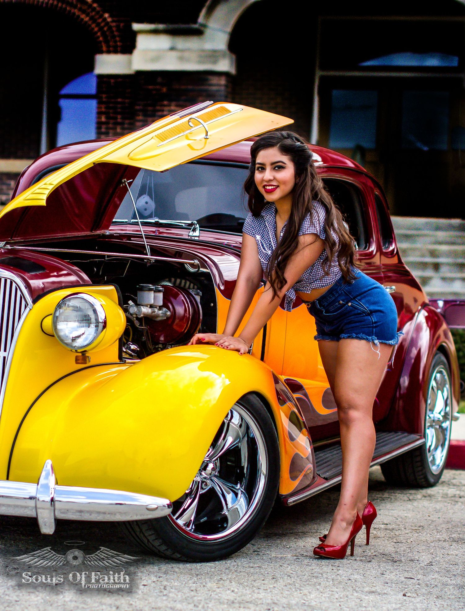 Woman in denim shorts poses beside a classic yellow and burgundy hot rod with flames at a car show.