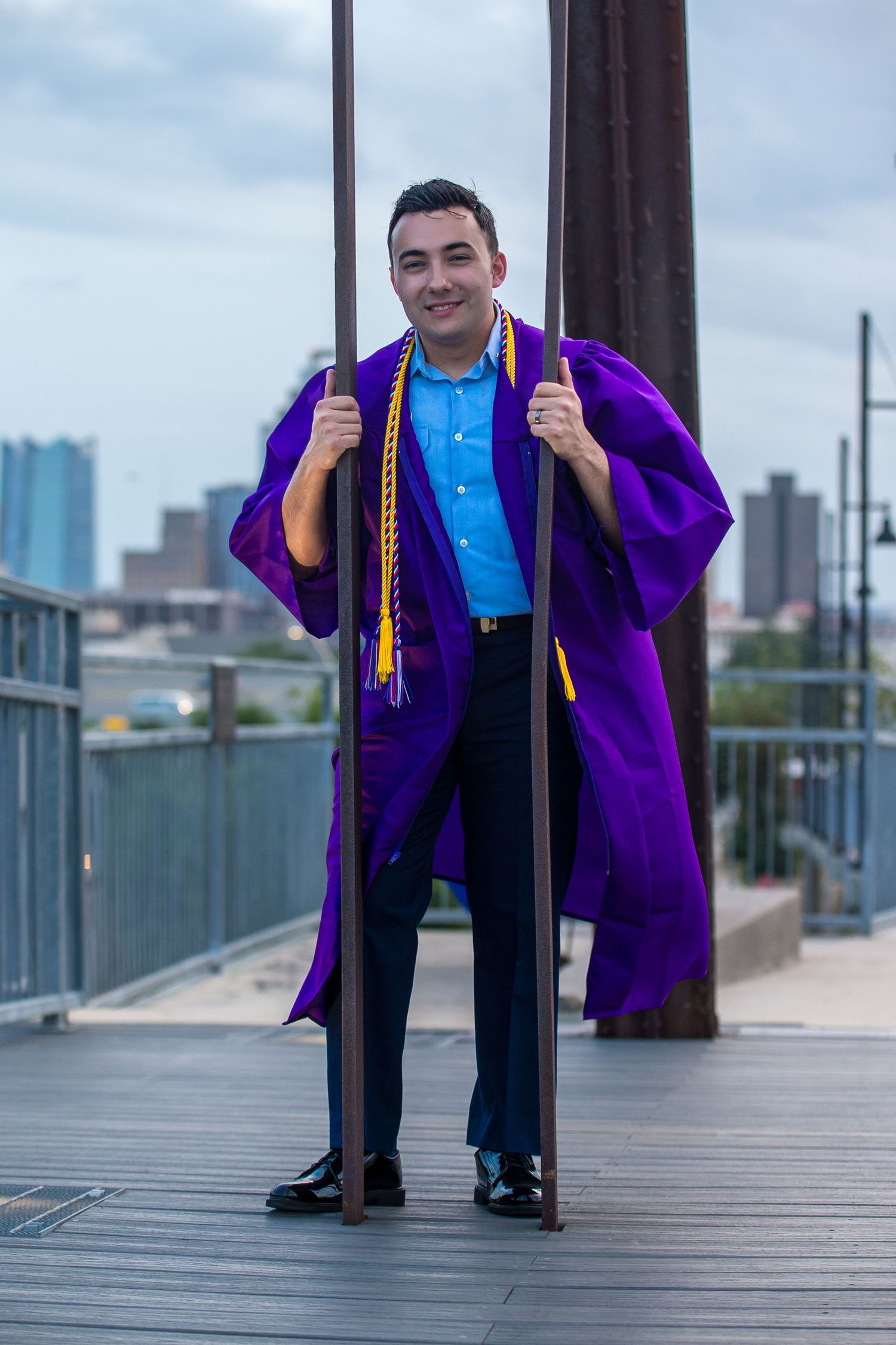 Young man in purple graduation gown and gold honor cords smiles proudly on a bridge with city skyline backdrop.