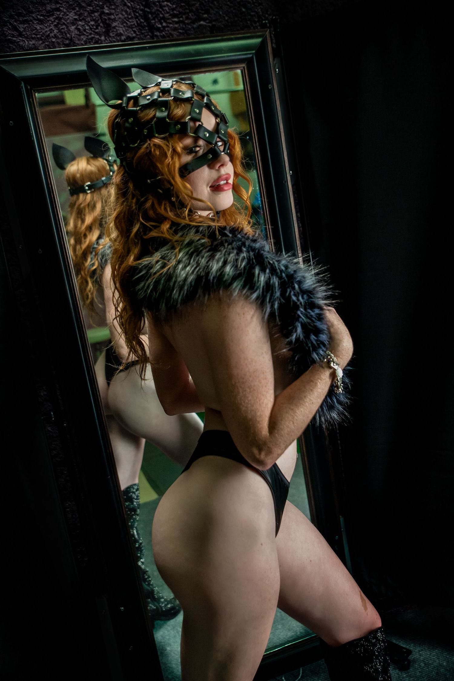 Red-haired woman in decorative mask and fur stole poses in black lingerie before a dark mirror.