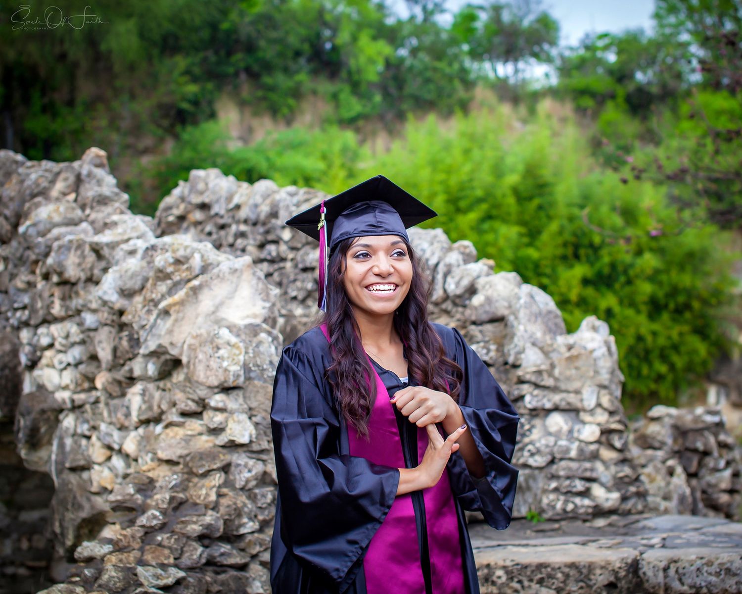 Smiling graduate in black cap and gown poses outdoors near stone ruins with lush green trees in background.