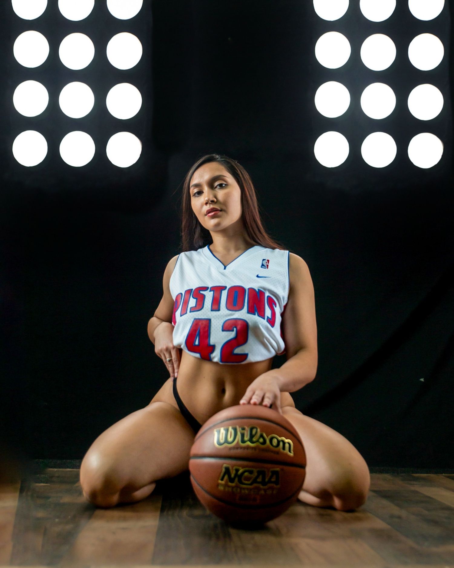 Woman in Detroit Pistons #42 jersey kneeling on court with Wilson NCAA basketball under studio lights.