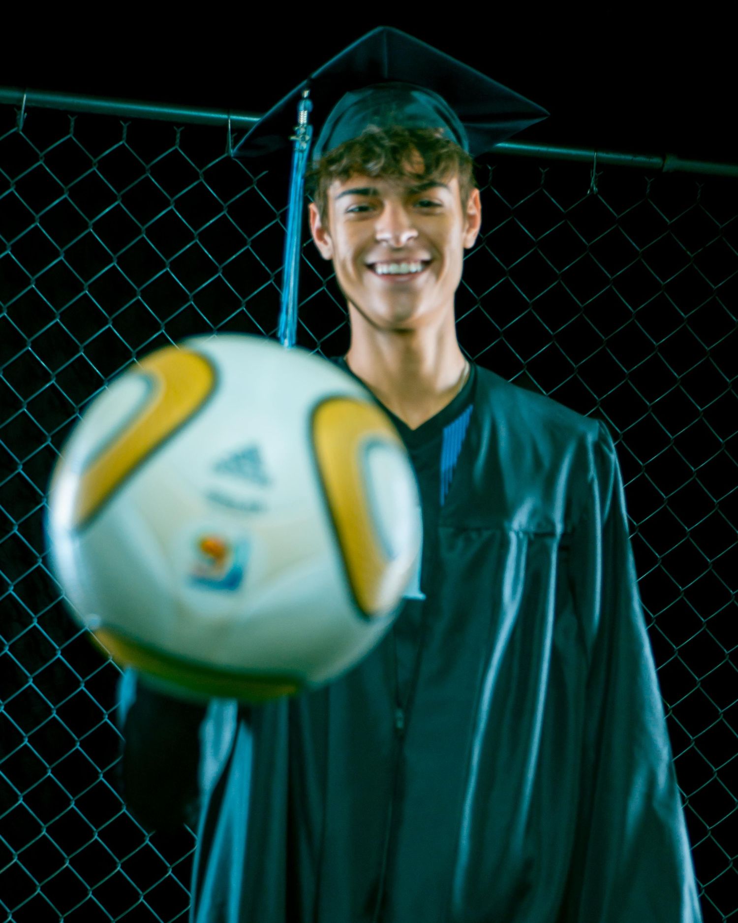 Smiling graduate in cap and gown holding a soccer ball in front of a chain-link fence at night.