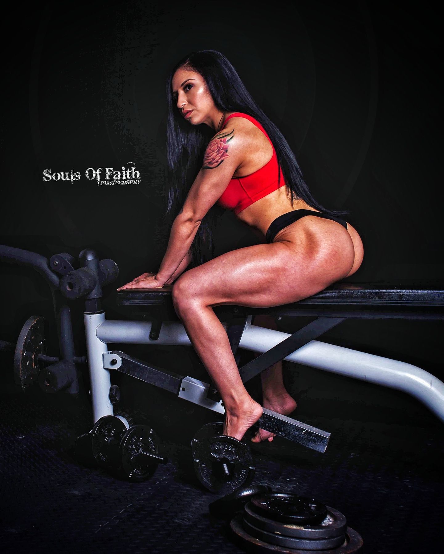 Muscular woman in red sports bra poses on gym bench surrounded by weights in dark studio setting.