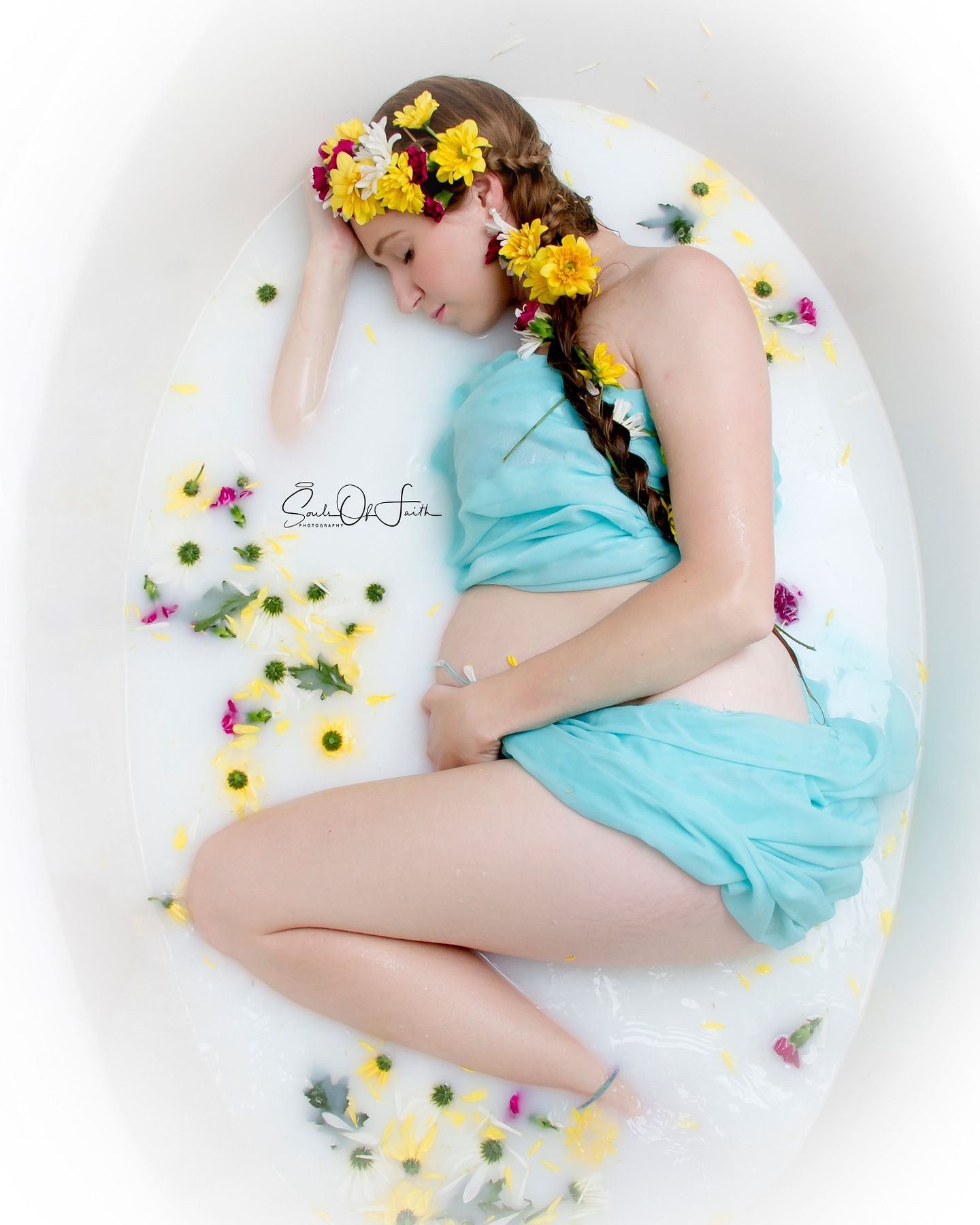 Pregnant woman in teal dress lying in floral milk bath with yellow and red flower crown.